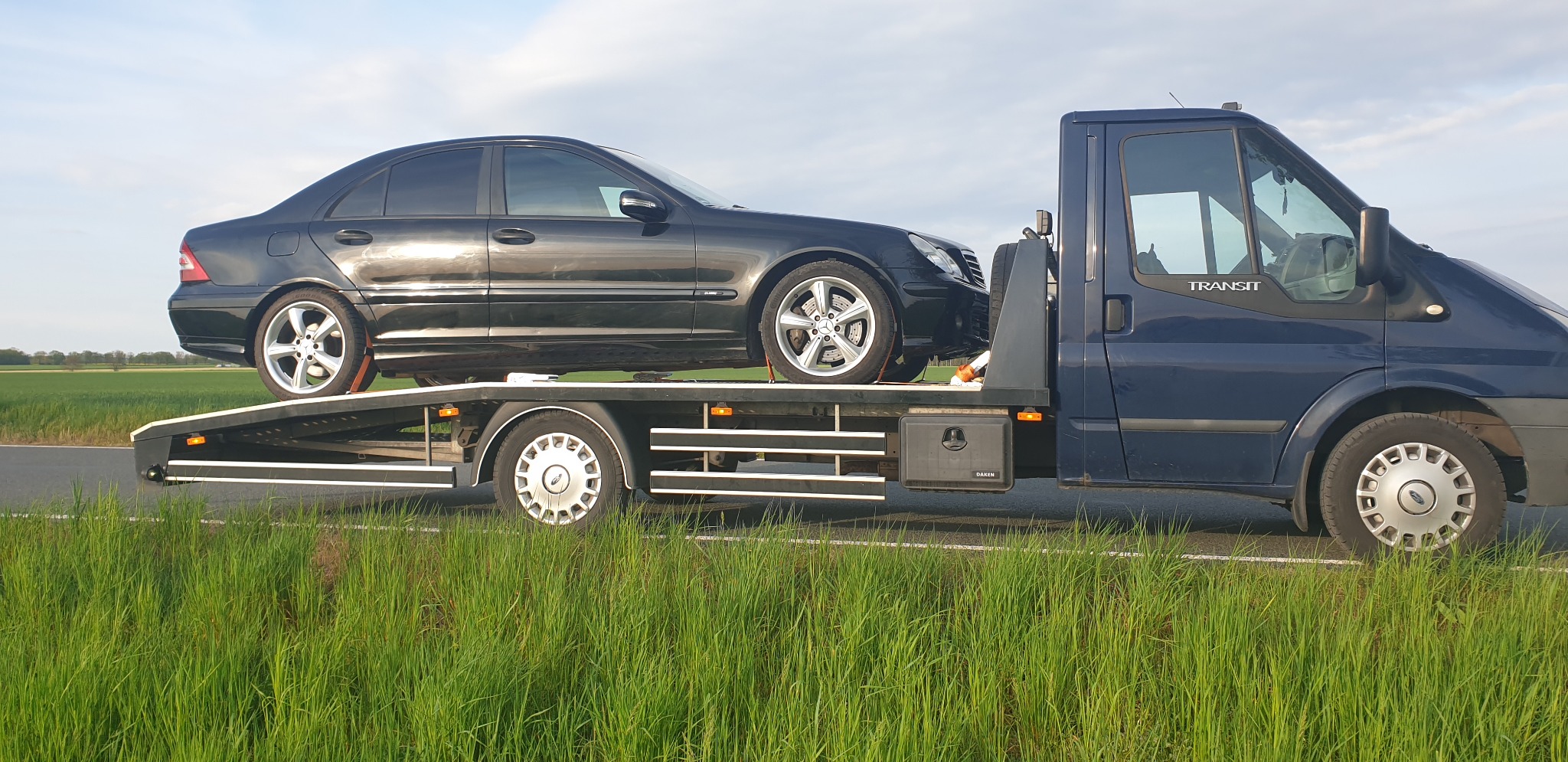 Czarny samochód osobowy marki Mercedes przewożony na lawecie Ford Transit w plenerze z zielonym polem w tle.