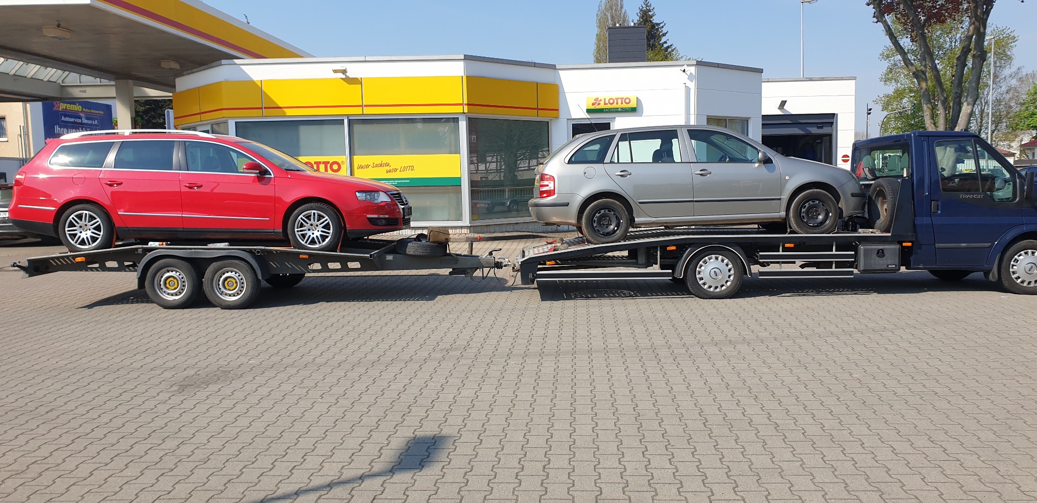 Dwa samochody osobowe, czerwony VW Passat i szary Skoda Fabia, załadowane na lawetę ciągniętą przez niebieskiego Forda Transita, na tle stacji benzynowej Shell i punktu Lotto.