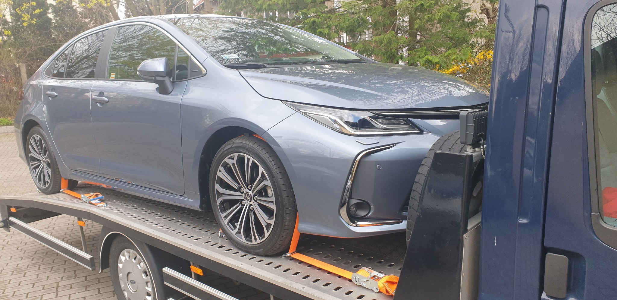 Szary samochód Toyota Corolla zabezpieczony pomarańczowymi pasami na platformie lawety, widok z boku, w tle drzewa.