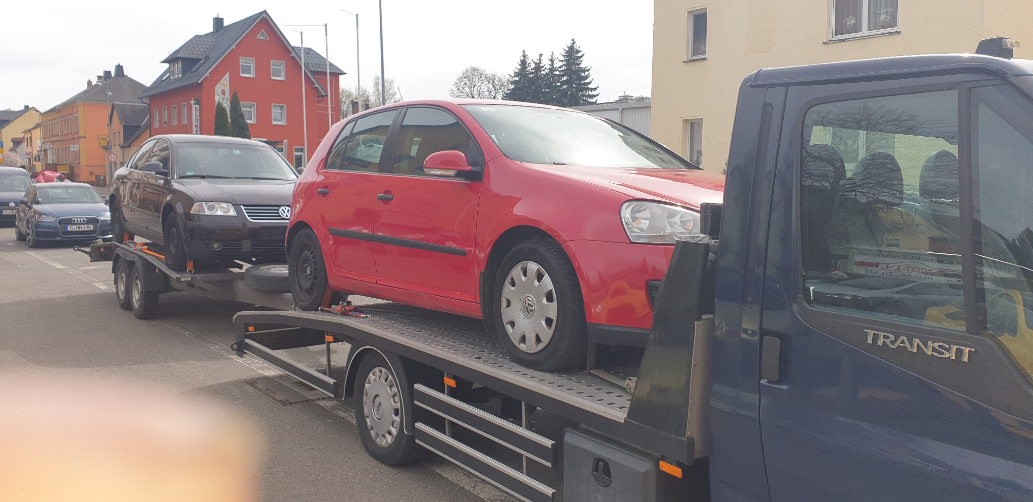Laweta przewozi czerwonego Volkswagena Golfa i ciemnego Volkswagena Passata na przyczepie w zabudowie miejskiej.