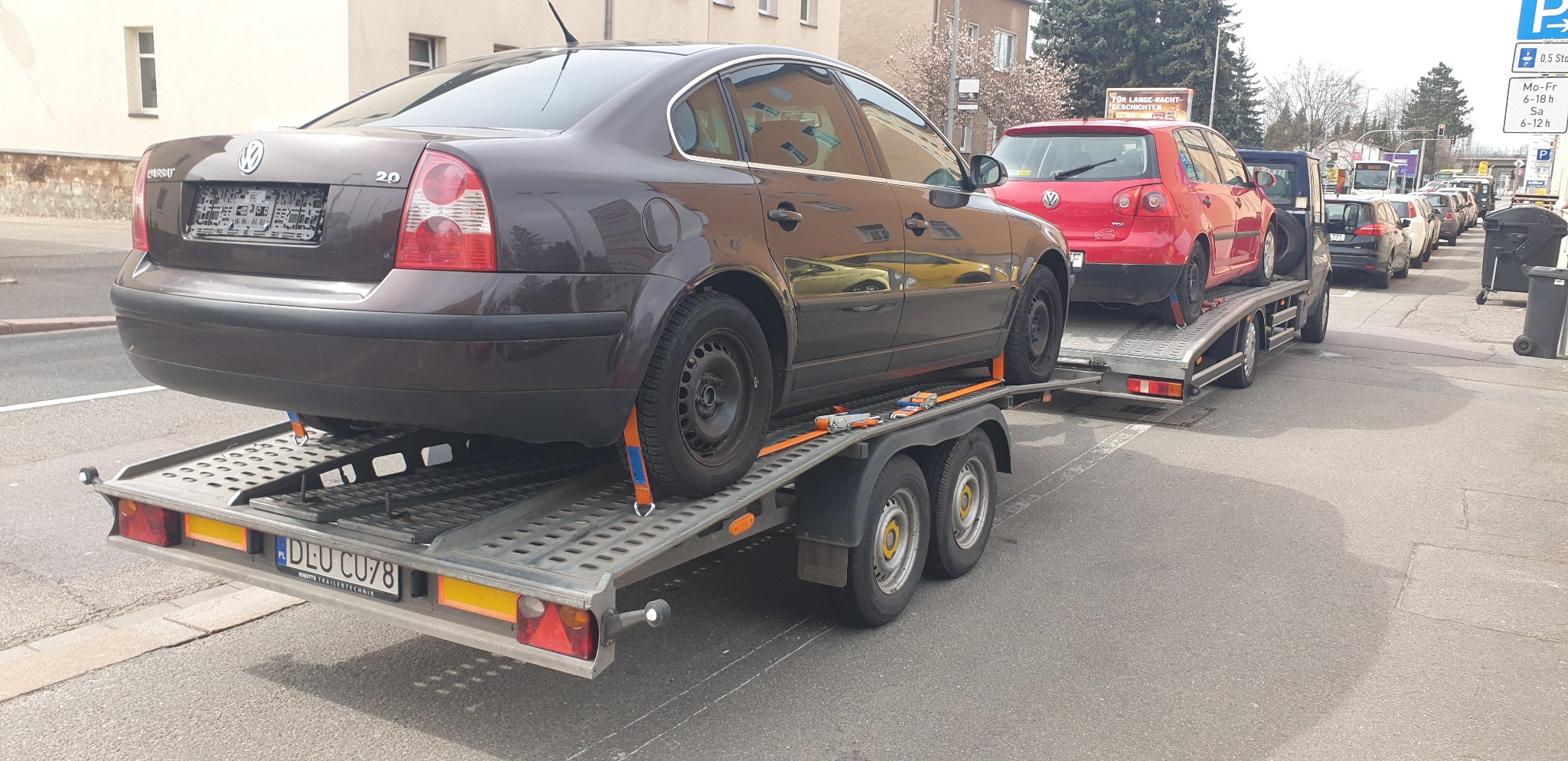 Brązowy Volkswagen Passat 2.0 zabezpieczony pomarańczowymi pasami na platformie lawety z polską rejestracją DLU CU78, za nim czerwony Volkswagen Golf, w tle ulica miejska z zaparkowanymi autami.