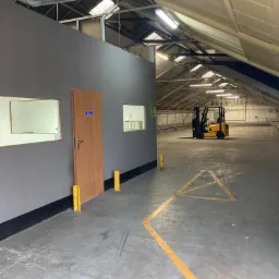 Wnętrze magazynu z betonową podłogą, żółtymi liniami wyznaczającymi ścieżki, ścianą z oknami i drzwiami oraz żółtym wózkiem widłowym w tle, oświetlonymi lampami sufitowymi.