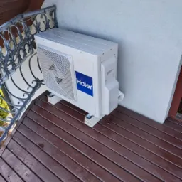 Zewnętrzna jednostka klimatyzacji Haier zamontowana na brązowym, drewnianym balkonie z ozdobną, metalową balustradą, przy białej ścianie budynku.