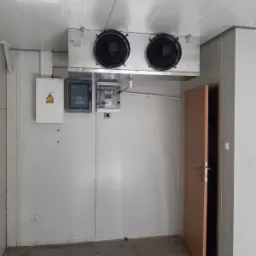 Wnętrze chłodni z agregatem chłodniczym podwieszonym pod sufitem, tablicami rozdzielczymi na ścianie i otwartymi drzwiami.