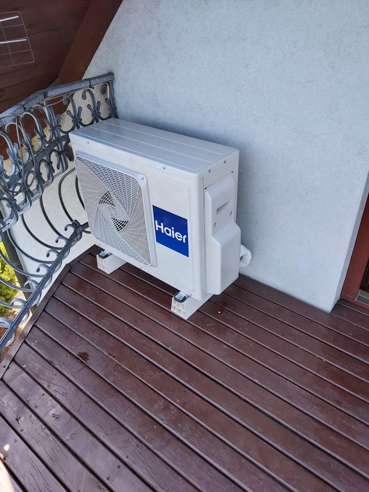 Zewnętrzna jednostka klimatyzacji Haier zamontowana na brązowym, drewnianym balkonie z ozdobną, metalową balustradą, przy białej ścianie budynku.