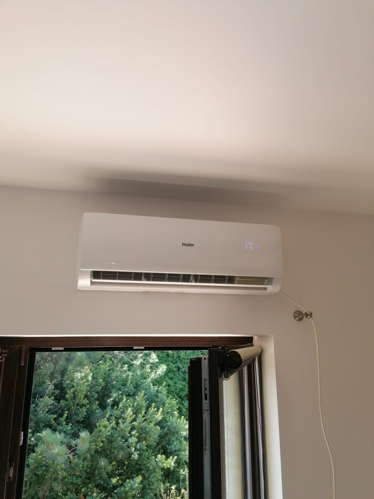 Biała jednostka klimatyzacji Haier zamontowana nad oknem z widokiem na zieleń, widoczny kabel zasilający przymocowany do ściany.