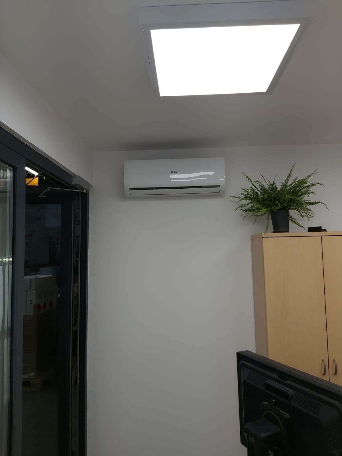 Biała jednostka wewnętrzna klimatyzacji Haier zamontowana na ścianie biura, obok szafki z paprotką i pod kwadratowym panelem LED w suficie.