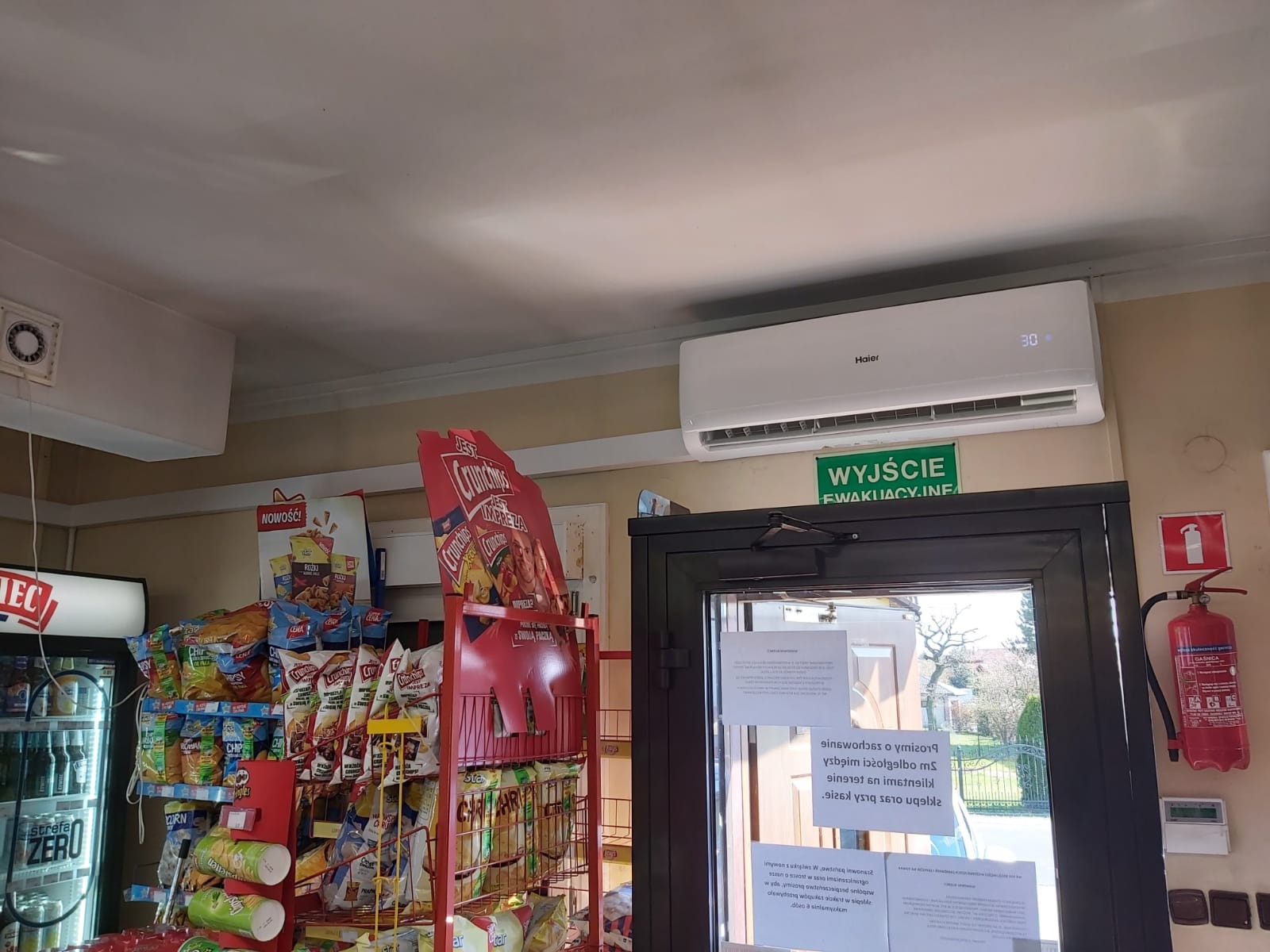 Biały klimatyzator Haier zamontowany w sklepie spożywczym, widoczny nad drzwiami ewakuacyjnymi i regałem z przekąskami.