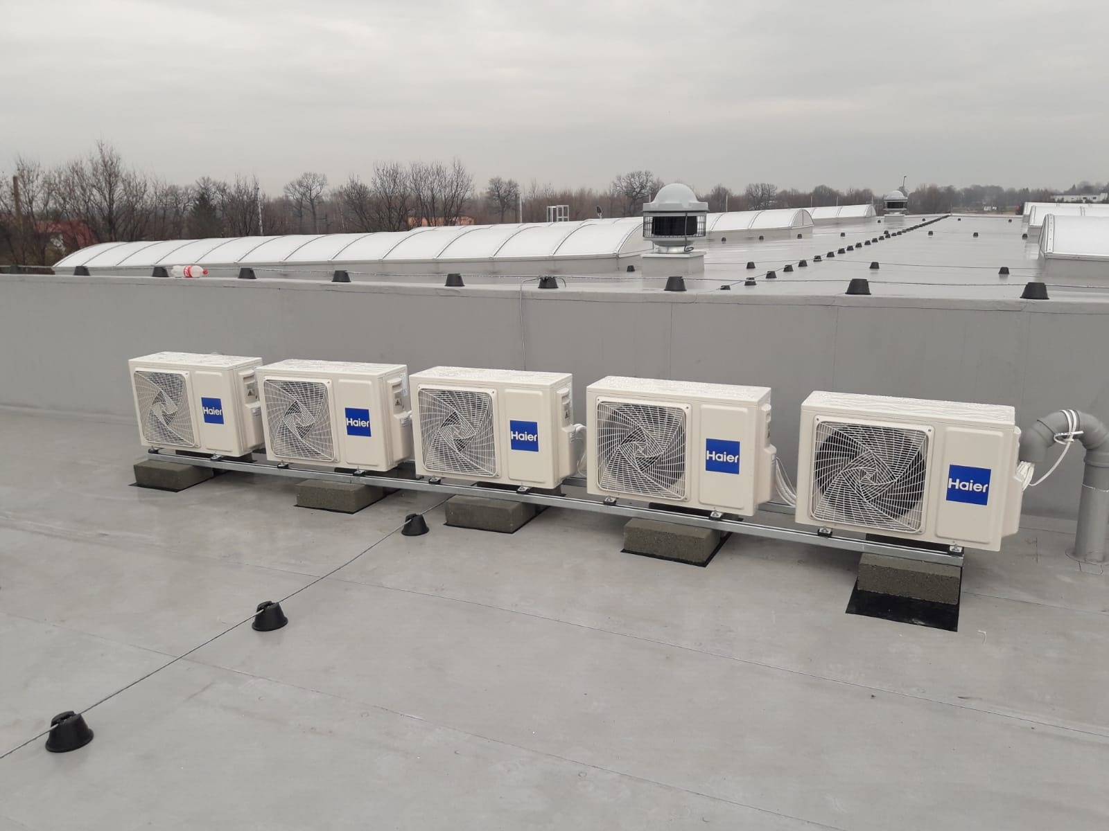 Rząd jednostek zewnętrznych klimatyzacji Haier na płaskim dachu budynku przemysłowego, zamontowanych na szarych podkładkach i metalowej szynie, z widocznymi przewodami rurowymi i elementami...