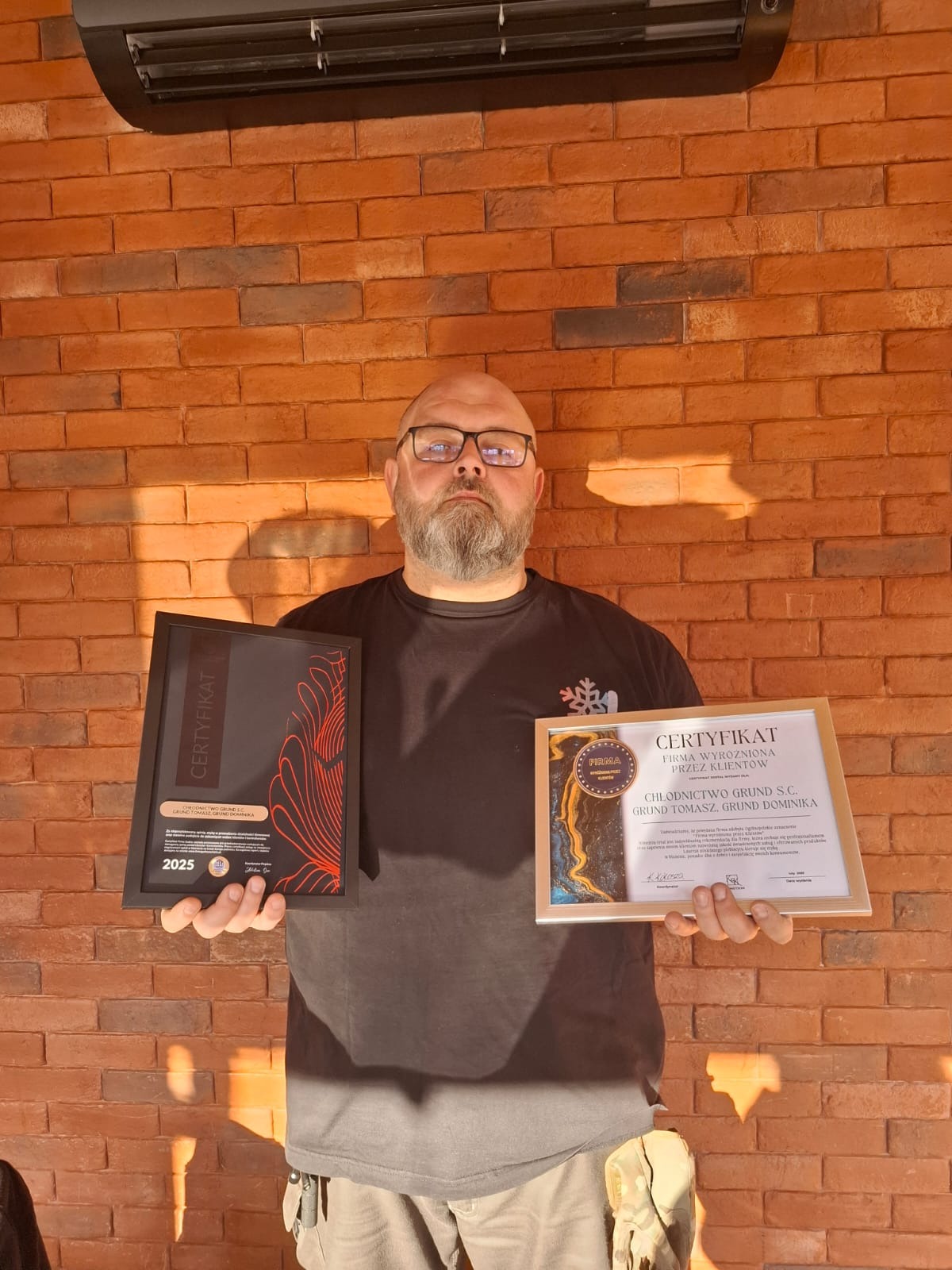 Mężczyzna w okularach prezentuje dwa certyfikaty firmy Chłodnictwo Grund S.C. na tle ceglanej ściany z zamontowanym klimatyzatorem.