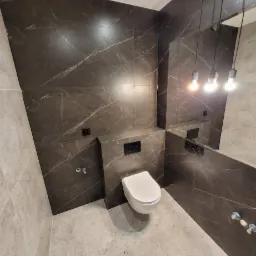 Wykończona łazienka z brązowymi, marmurkowymi płytkami na ścianach i jasnymi płytkami na podłodze. Wisząca toaleta i minimalistyczne oświetlenie.