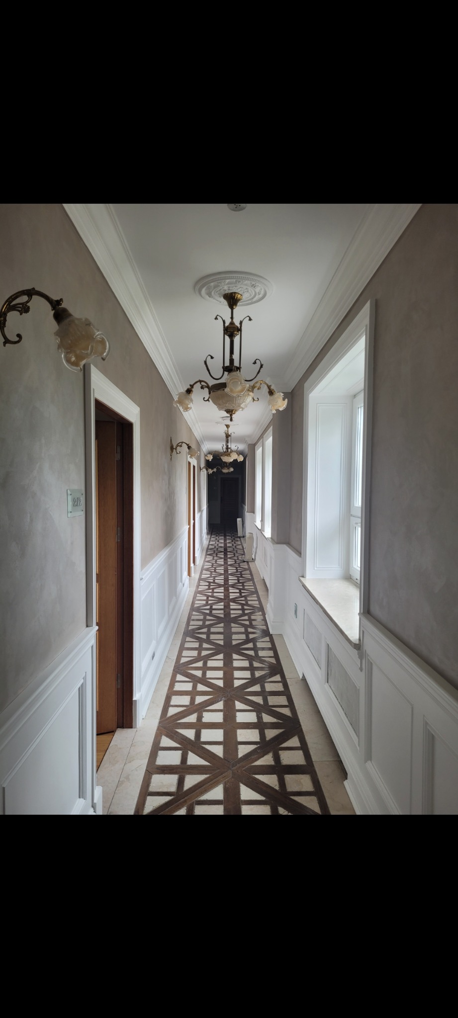 Długi korytarz po remoncie z wzorzystą podłogą, kremowymi ścianami z boazerią i ozdobnymi lampami wiszącymi. Widok w głąb korytarza.