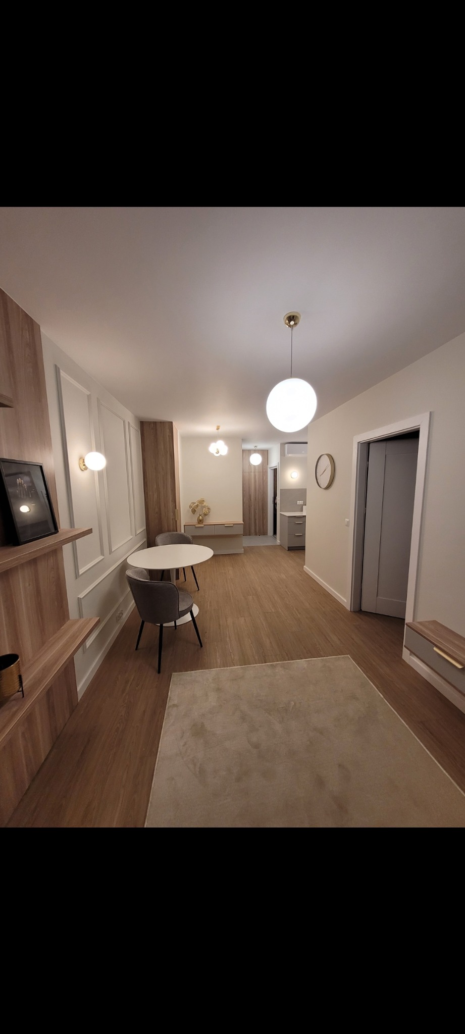 Jasne wnętrze po remoncie: drewniana podłoga, białe ściany z dekoracyjnymi panelami, okrągły stół i nowoczesne oświetlenie kuliste. Widok na kuchnię w tle.