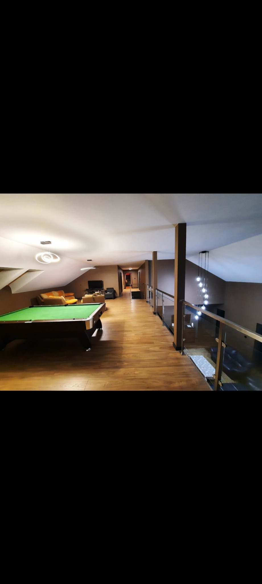 Podłużny pokój na poddaszu z parkietem, stołem bilardowym i szklaną balustradą. Widoczne oświetlenie i meble. Ukończony projekt aranżacji wnętrza.
