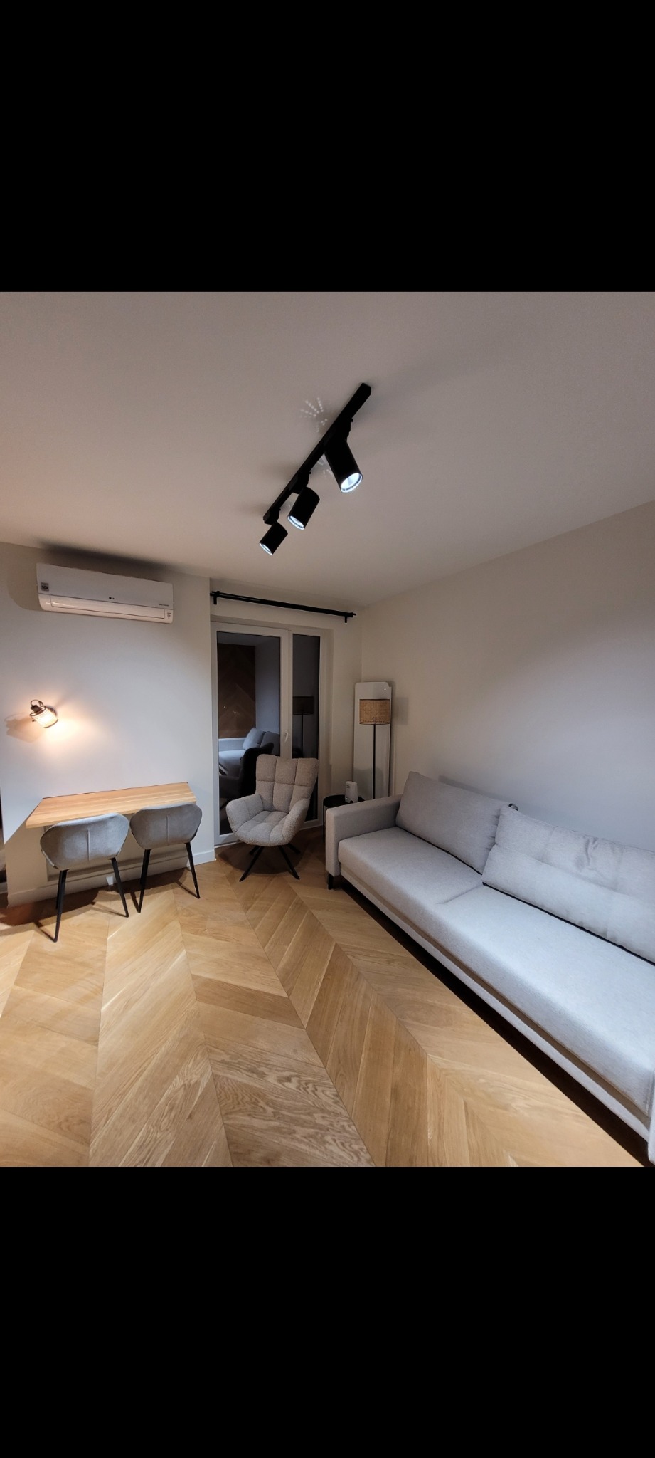 Salon z parkietem w jodełkę: beżowa sofa, szare krzesła przy biurku i fotel. Oświetlenie sufitowe punktowe. Widok na balkon. Minimalistyczny design.