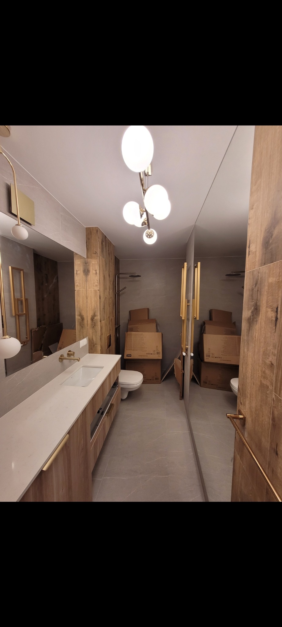 Wykończona łazienka z drewnianymi akcentami, dużym lustrem i nowoczesnym oświetleniem. Widoczne kartony sugerują etap po remoncie lub tuż przed wprowadzeniem.