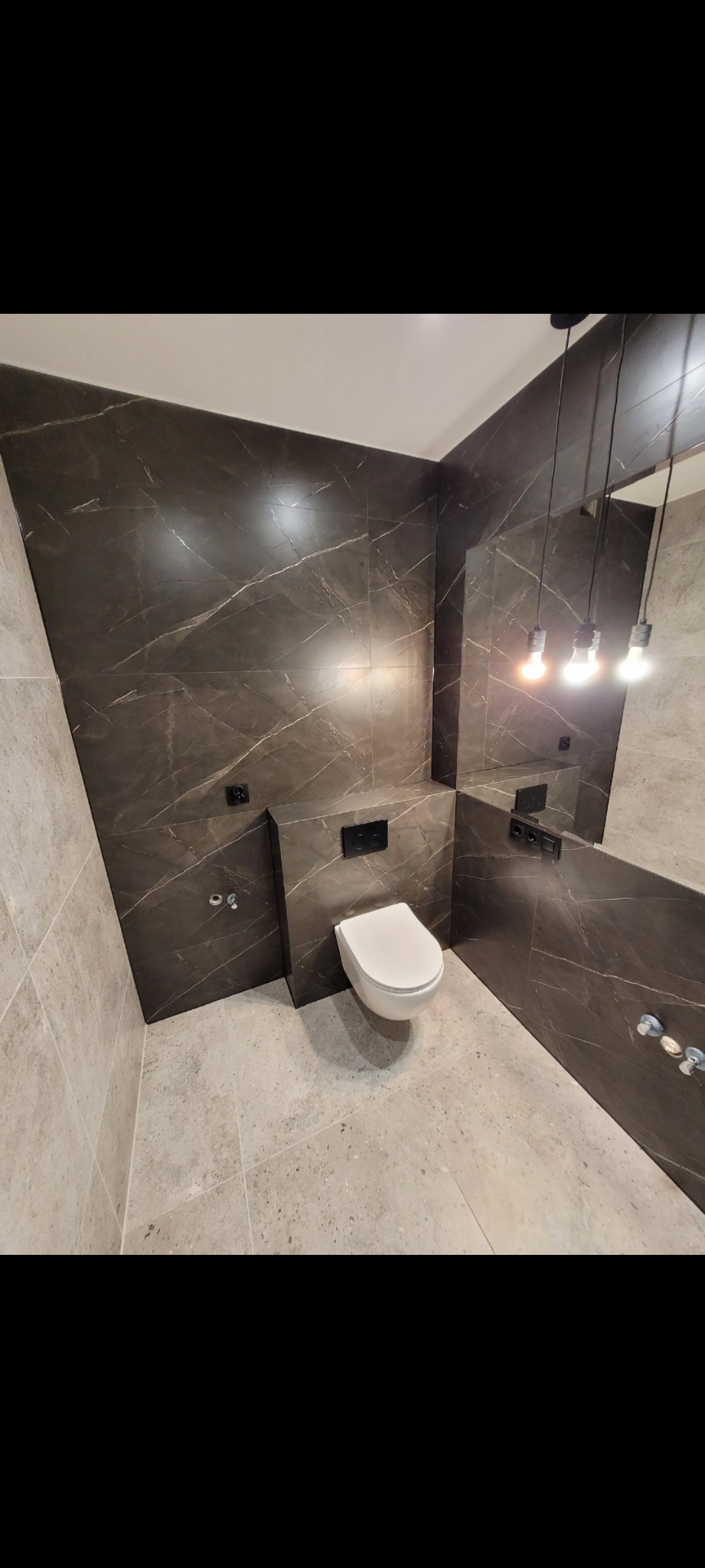 Wykończona łazienka z brązowymi, marmurkowymi płytkami na ścianach i jasnymi płytkami na podłodze. Wisząca toaleta i minimalistyczne oświetlenie.