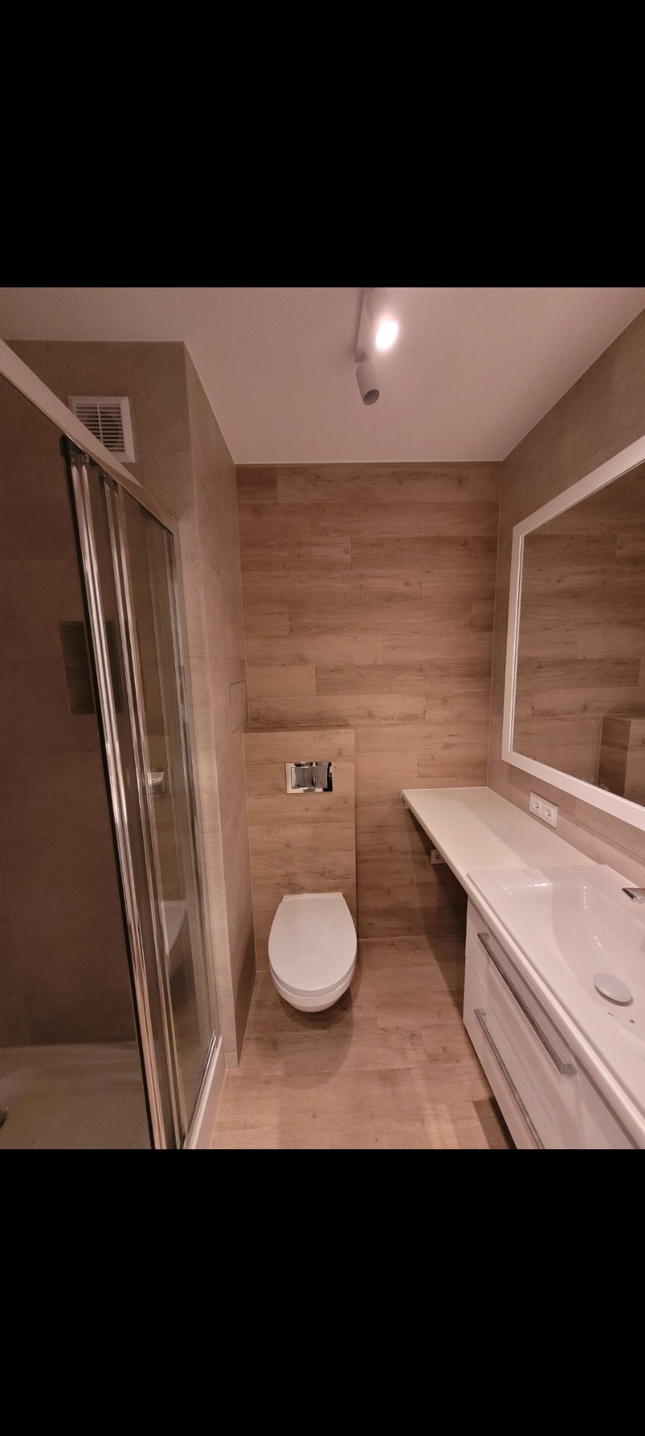 Nowoczesna łazienka z kabiną prysznicową, podwieszaną toaletą i umywalką z szafką, wykończona płytkami imitującymi drewno, oświetlona punktowo.