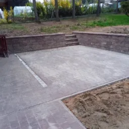 Wyłożony kostką plac z murem oporowym, widoczne schodki w murze, odpływ liniowy i fragment terenu z ziemią.