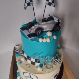 Dwupoziomowy tort urodzinowy z motywem Formuły 1: turkusowy górny poziom z modelem srebrnego bolidu i flagami w szachownicę, dolny poziom w kolorze kremowym z logo Mercedesa i dekoracjami...
