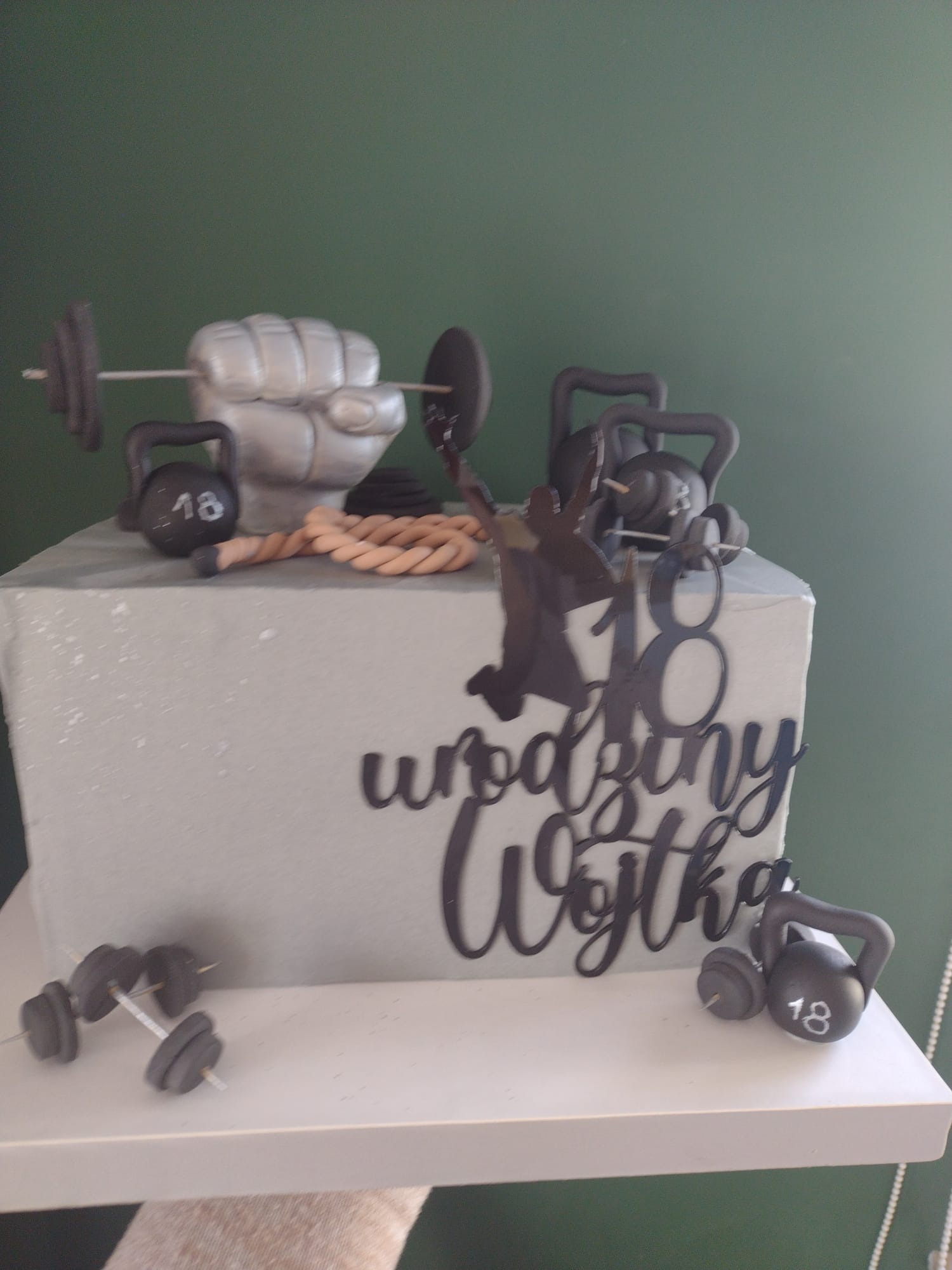 Tort urodzinowy w stylu fitness z dekoracjami: sztanga w dłoni, kettlebells z numerem 18, lina i napis 'Urodziny Wojtka'.