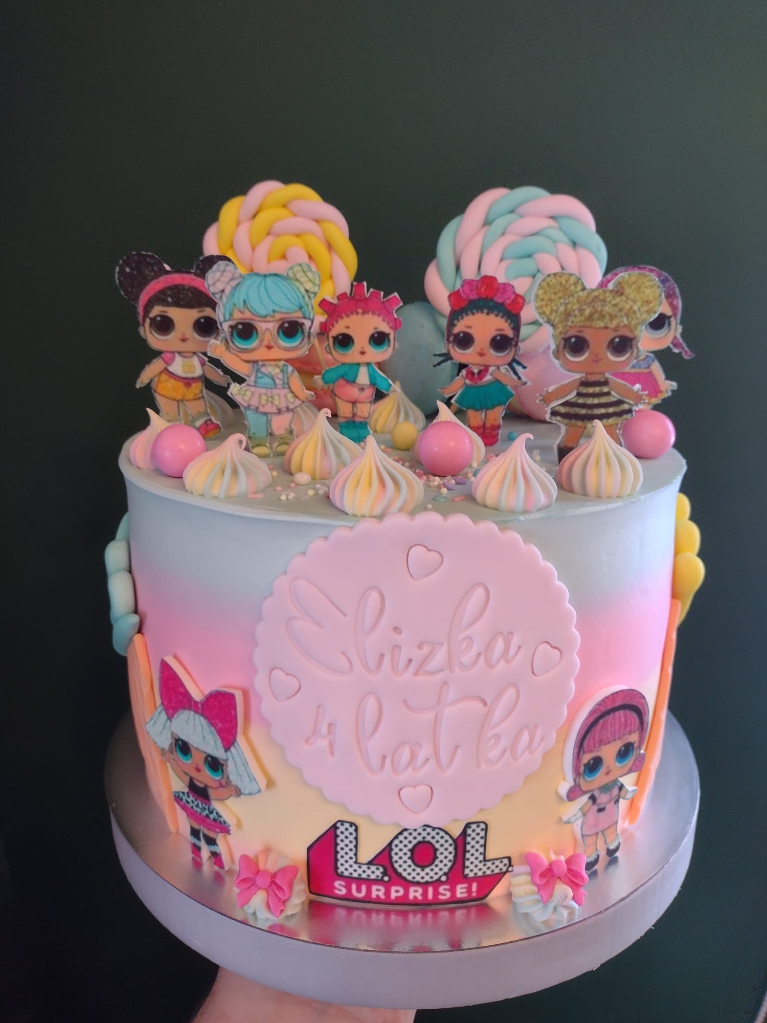 Tort urodzinowy dla dziewczynki z motywem LOL Surprise, ozdobiony lizakami z masy cukrowej i figurkami postaci na górze, z napisem 'Elizka 4 latka' na różowym tle.