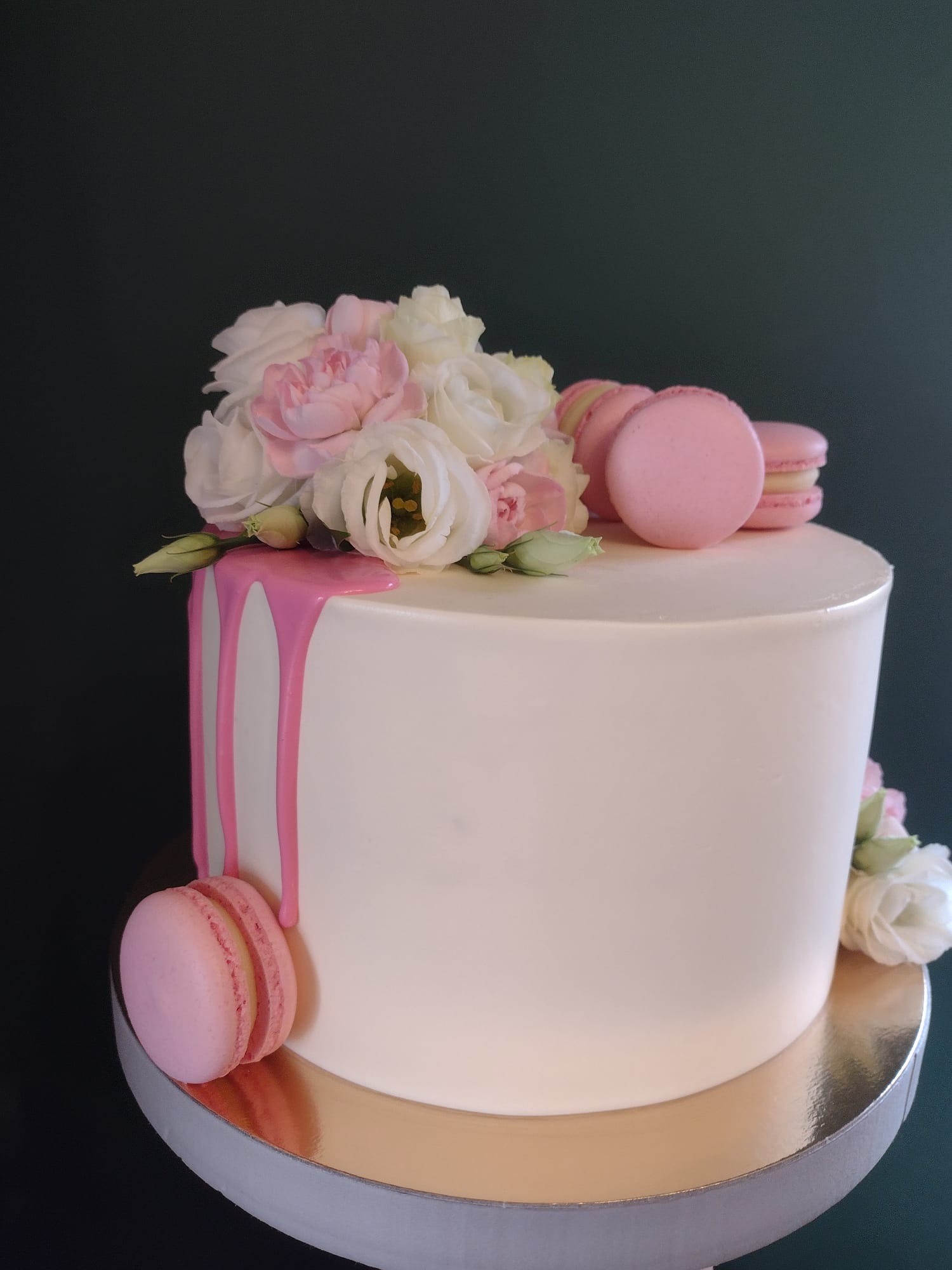 Elegancki tort z białym kremem, różowym lukrem spływającym po boku, udekorowany białymi i różowymi kwiatami oraz różowymi makaronikami, umieszczony na złotej paterze.