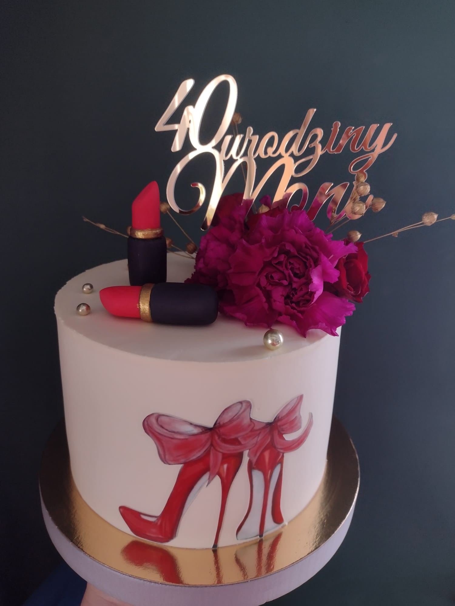 Tort urodzinowy z dekoracją w stylu glamour: złoty napis '40 Urodziny Moni', kwiaty, pomadki i rysunek szpilek.