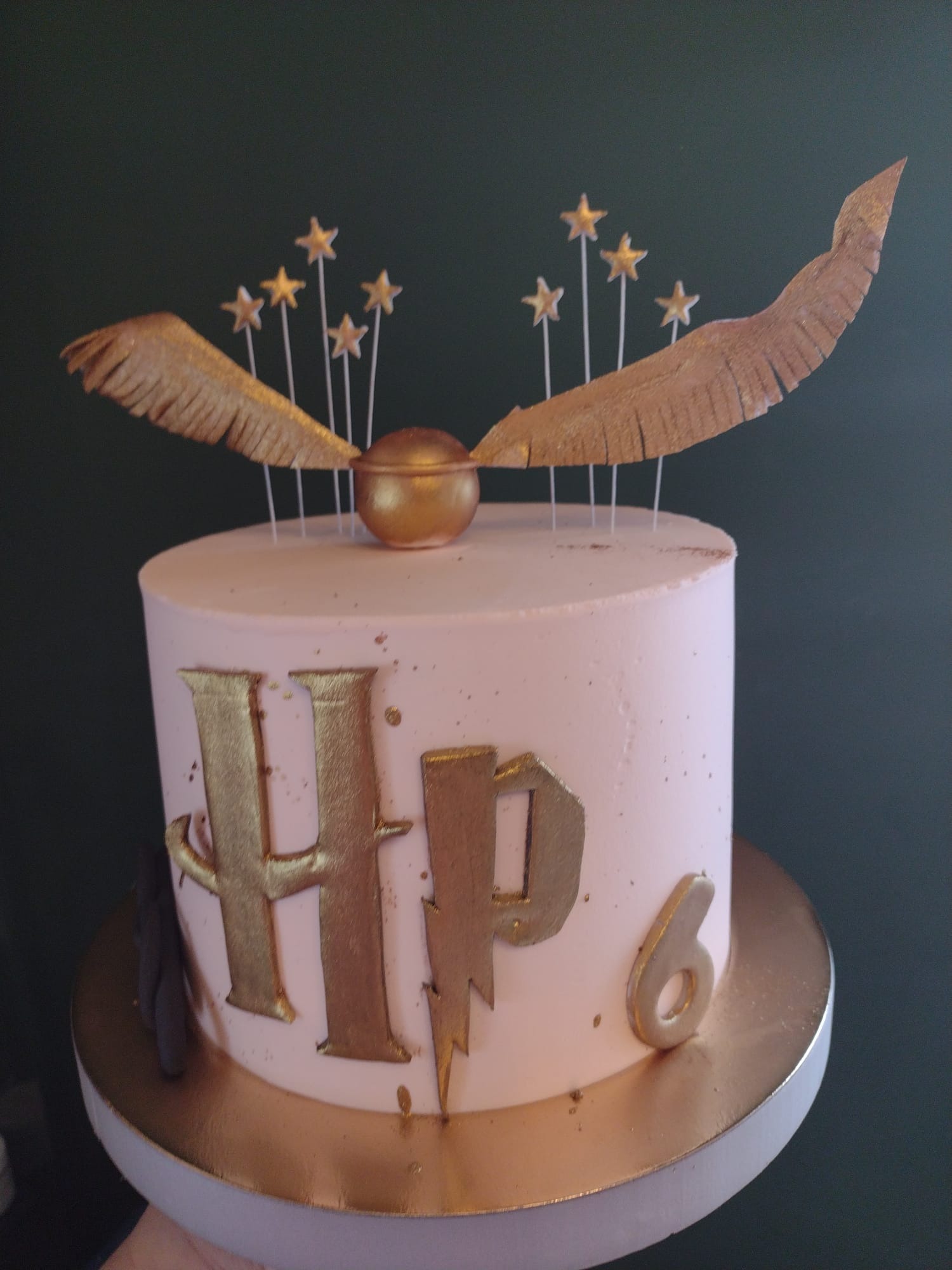 Tort urodzinowy w jasnoróżowym kremie, zdobiony złotymi elementami nawiązującymi do Harry'ego Pottera: inicjały HP, błyskawica, złoty znicz ze skrzydłami i gwiazdki. Na torcie widoczna cyfra 6.