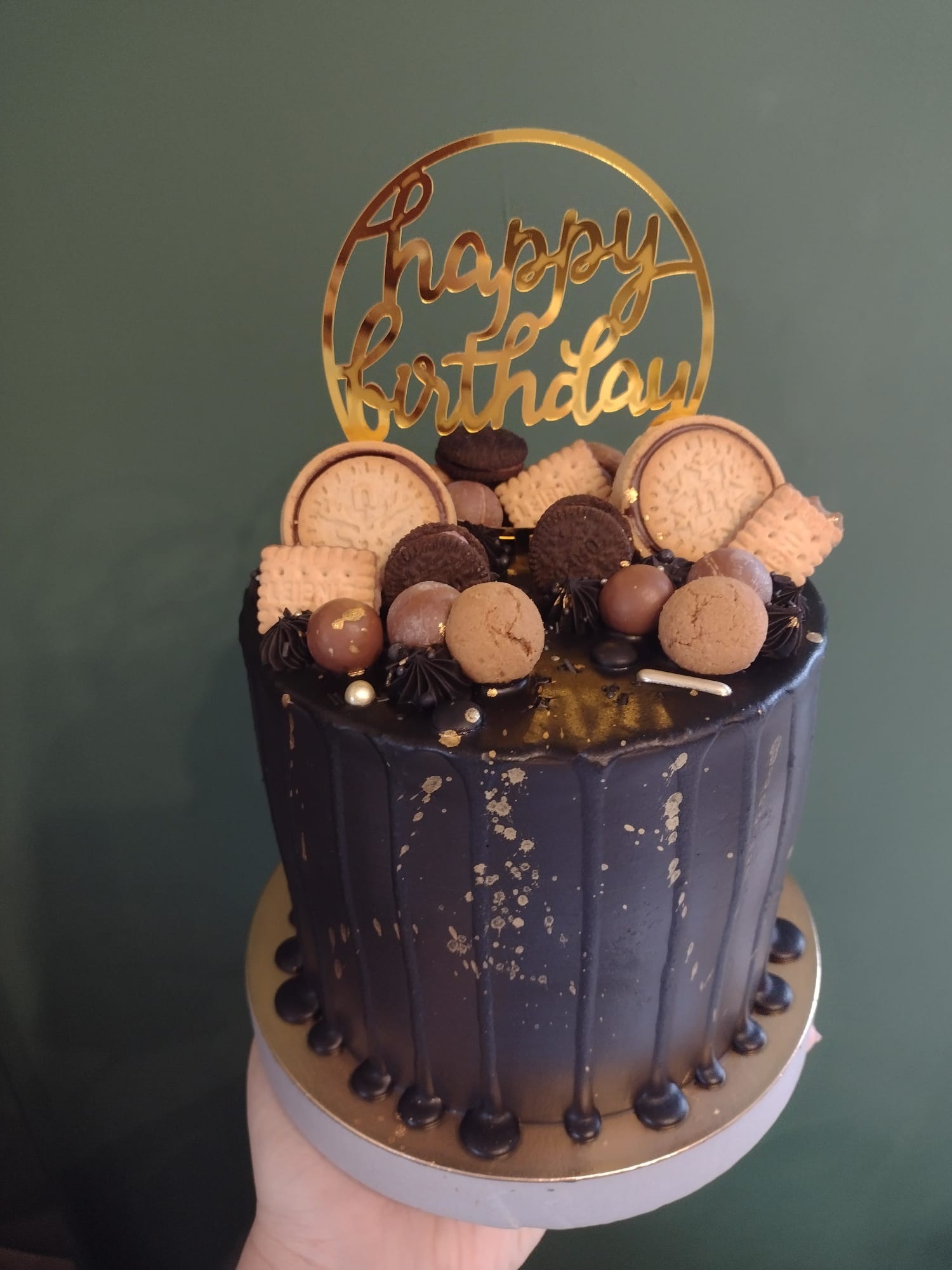 Ciemny tort urodzinowy z złotym napisem, dekorowany ciastkami, kuleczkami i złotymi plamkami, trzymany w dłoni.
