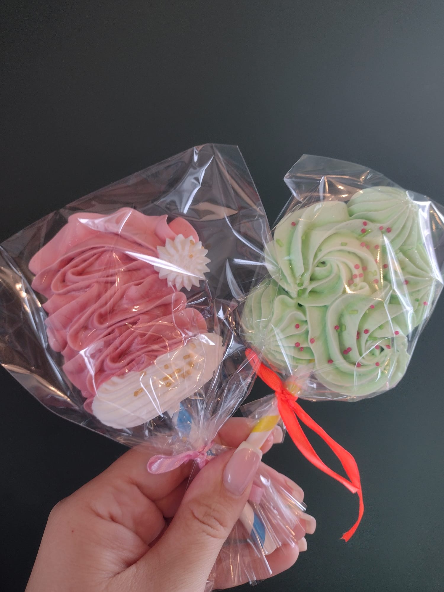 Dwa zapakowane lizaki bezowe: różowy z białą dekoracją kwiatową i złotymi posypkami oraz zielony z czerwonymi posypkami, trzymane w dłoni na tle ciemnej powierzchni.