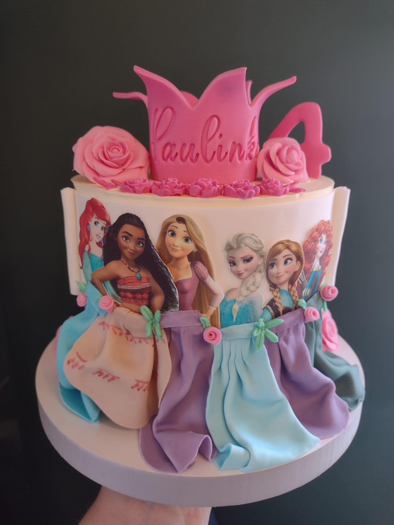 Dwupoziomowy tort urodzinowy z motywem księżniczek Disneya, zdobiony różową koroną z napisem 'Paulina' i cyfrą '4', otoczony różami z lukru.