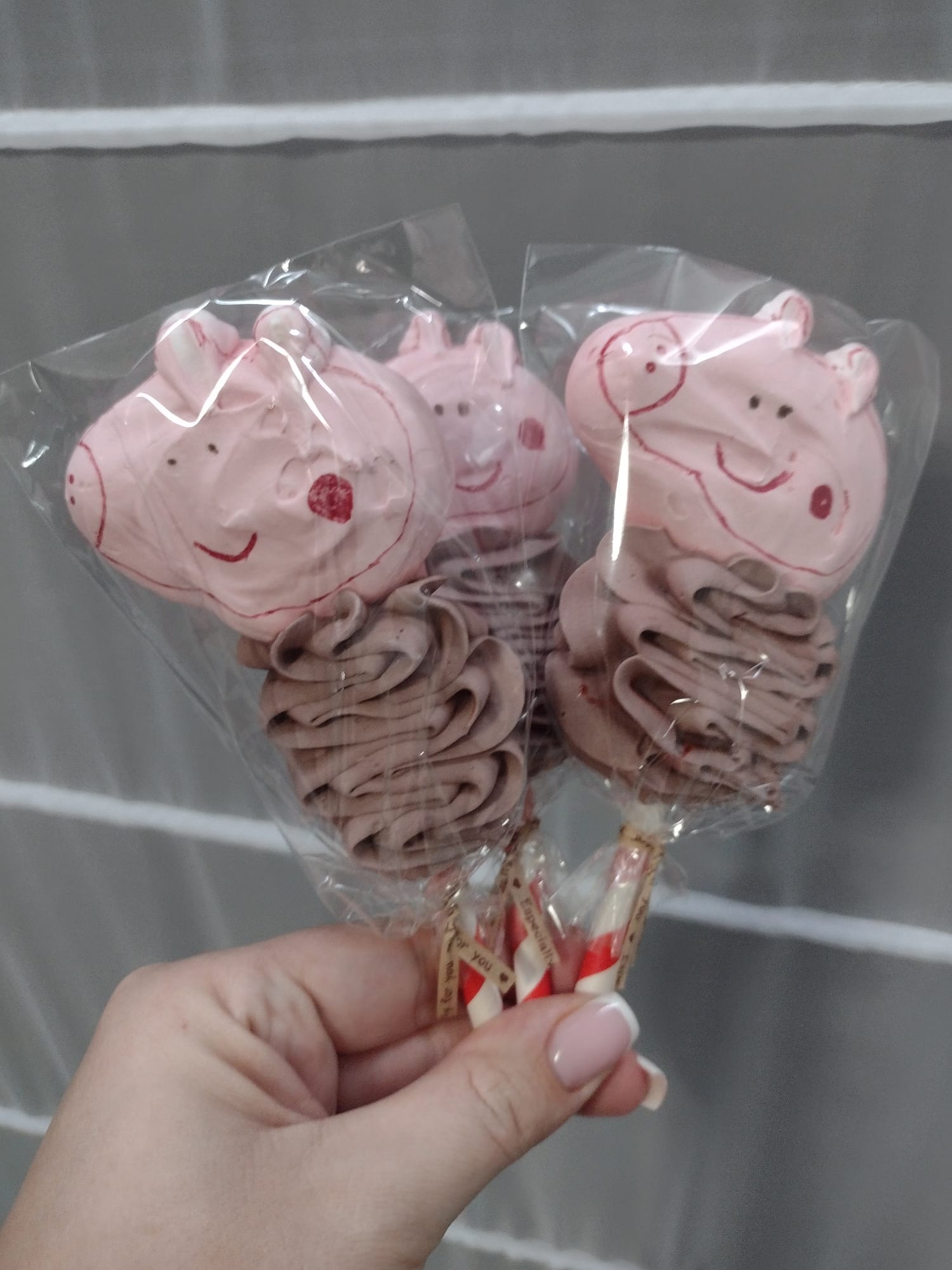 Ręka trzyma trzy lizaki bezowe w kształcie świnki, zapakowane w przezroczysty celofan, z brązową bezą u dołu i różową głową świnki na górze, na patyku z dekoracyjną wstążką.