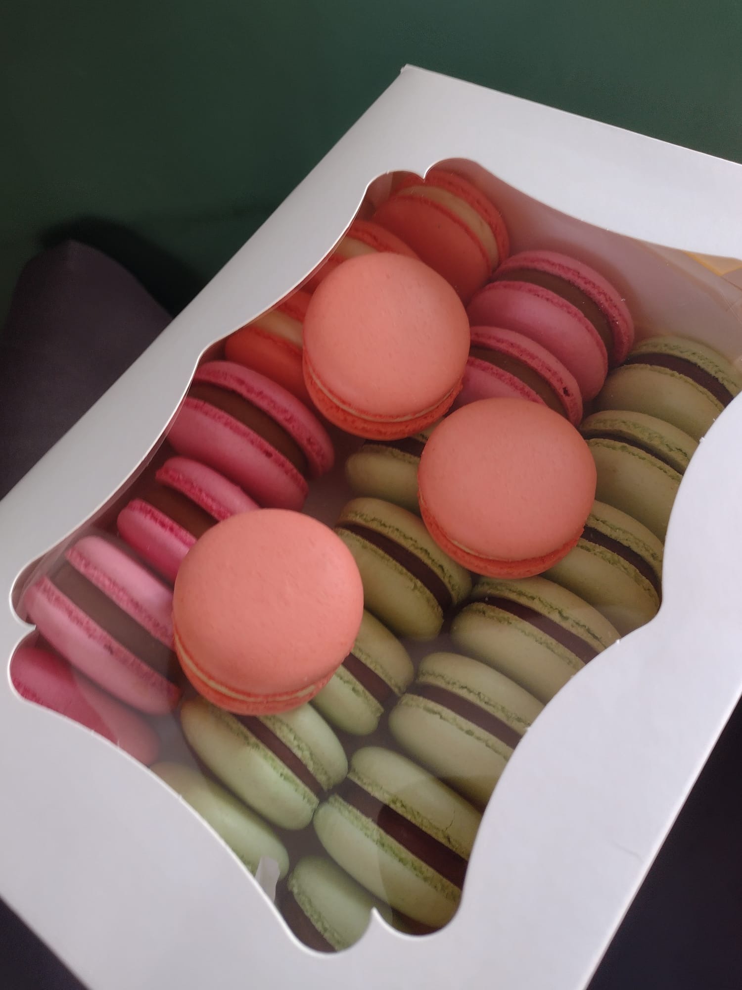 Kartonowe pudełko z makaronikami w pastelowych kolorach: łososiowe, różowe i pistacjowe, z czekoladowym kremem.