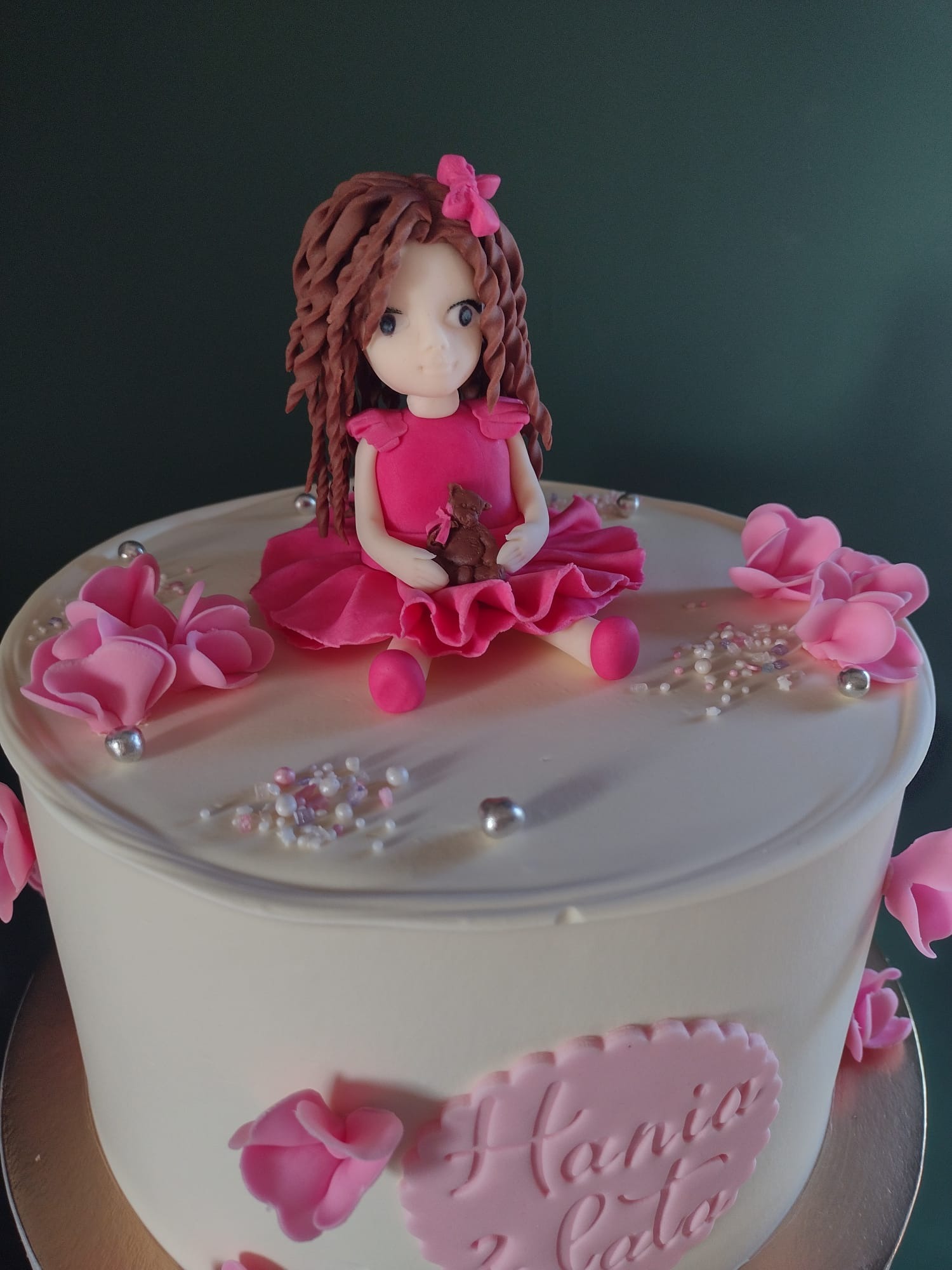 Tort urodzinowy z figurką dziewczynki w różowej sukience trzymającej misia, ozdobiony różowymi kwiatami i srebrnymi kuleczkami, wykonany w Wilczy.