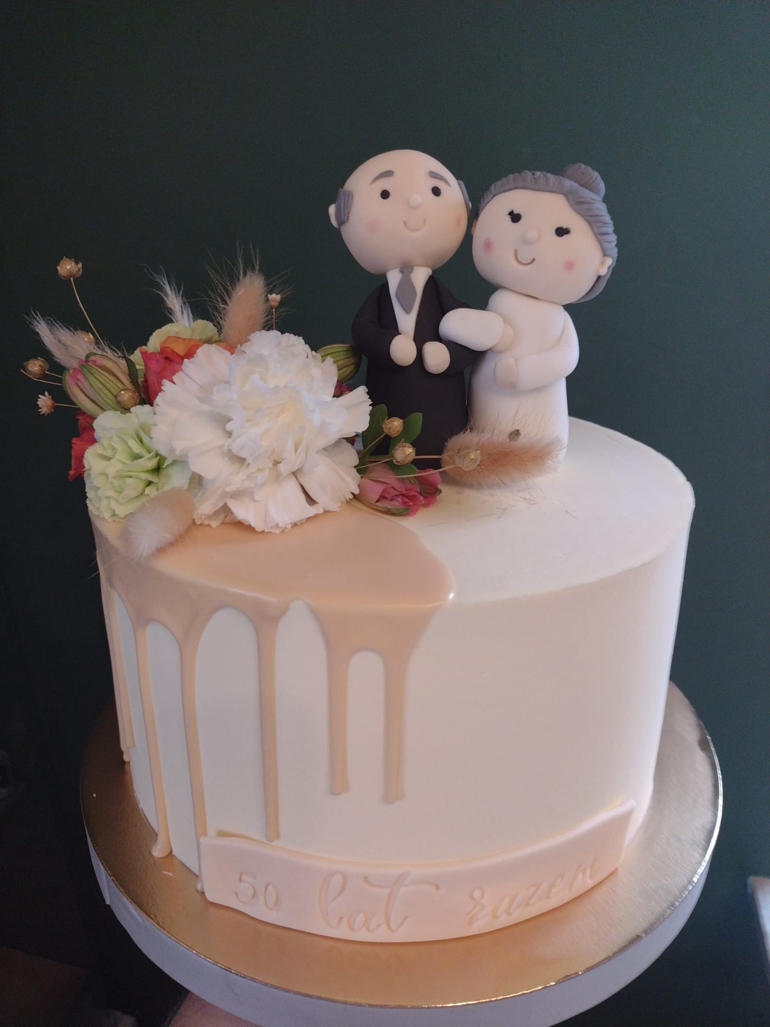 Tort okolicznościowy z figurkami pary starszych osób, dekorowany kwiatami i napisem '50 lat razem', z polewą w odcieniach beżu.