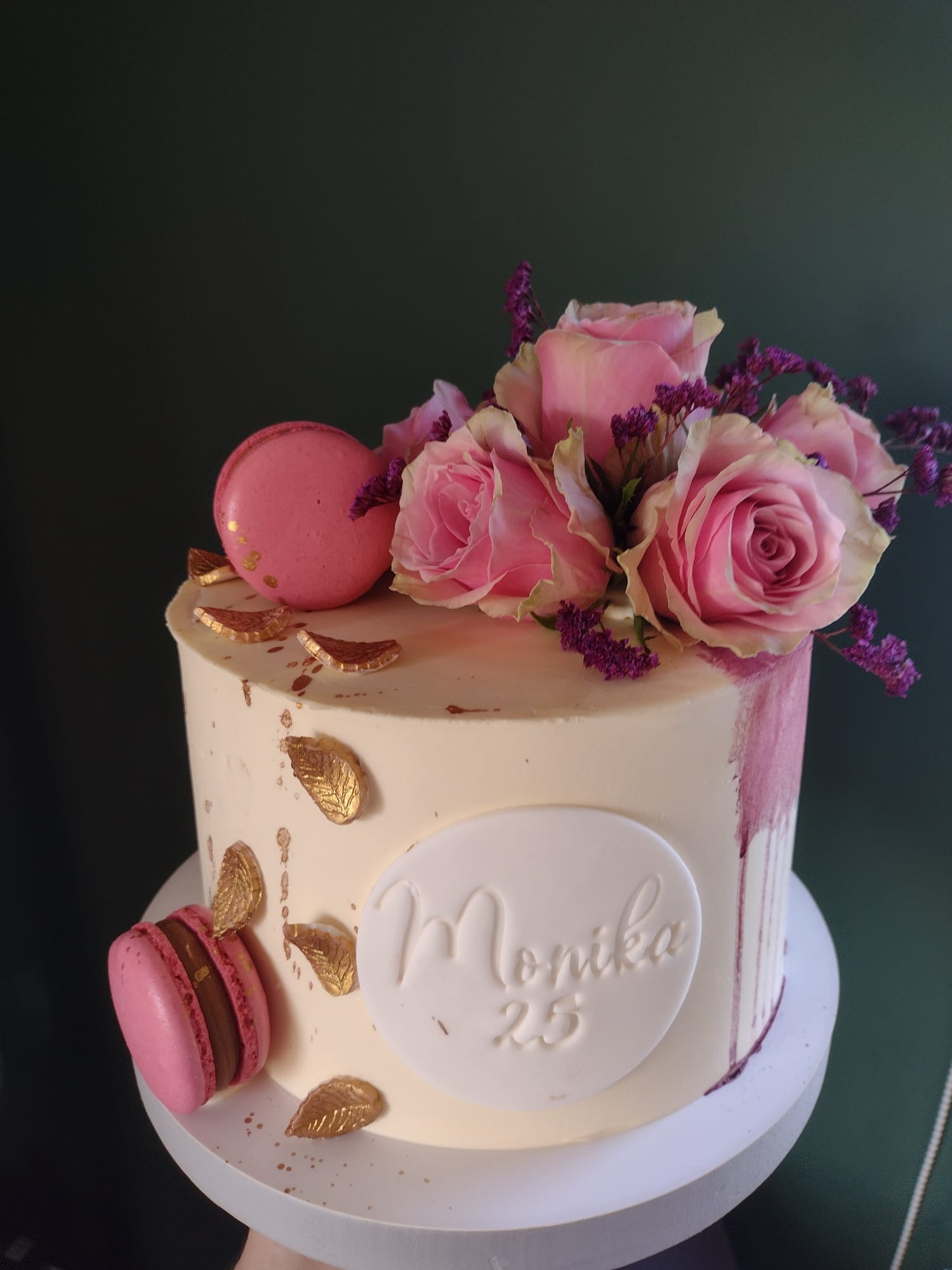 Tort urodzinowy z białym kremem, różowymi różami i makaronikami, dekorowany złotymi listkami i napisem 'Monika 25'.