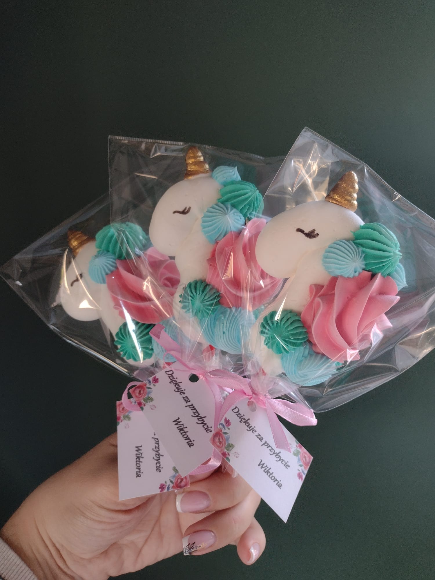 Bukiet trzech zapakowanych w folię bezowych jednorożców z różowymi i turkusowymi dekoracjami, przywiązanych różową wstążką z bilecikami z podziękowaniem trzymany w dłoni z delikatnym manicure.