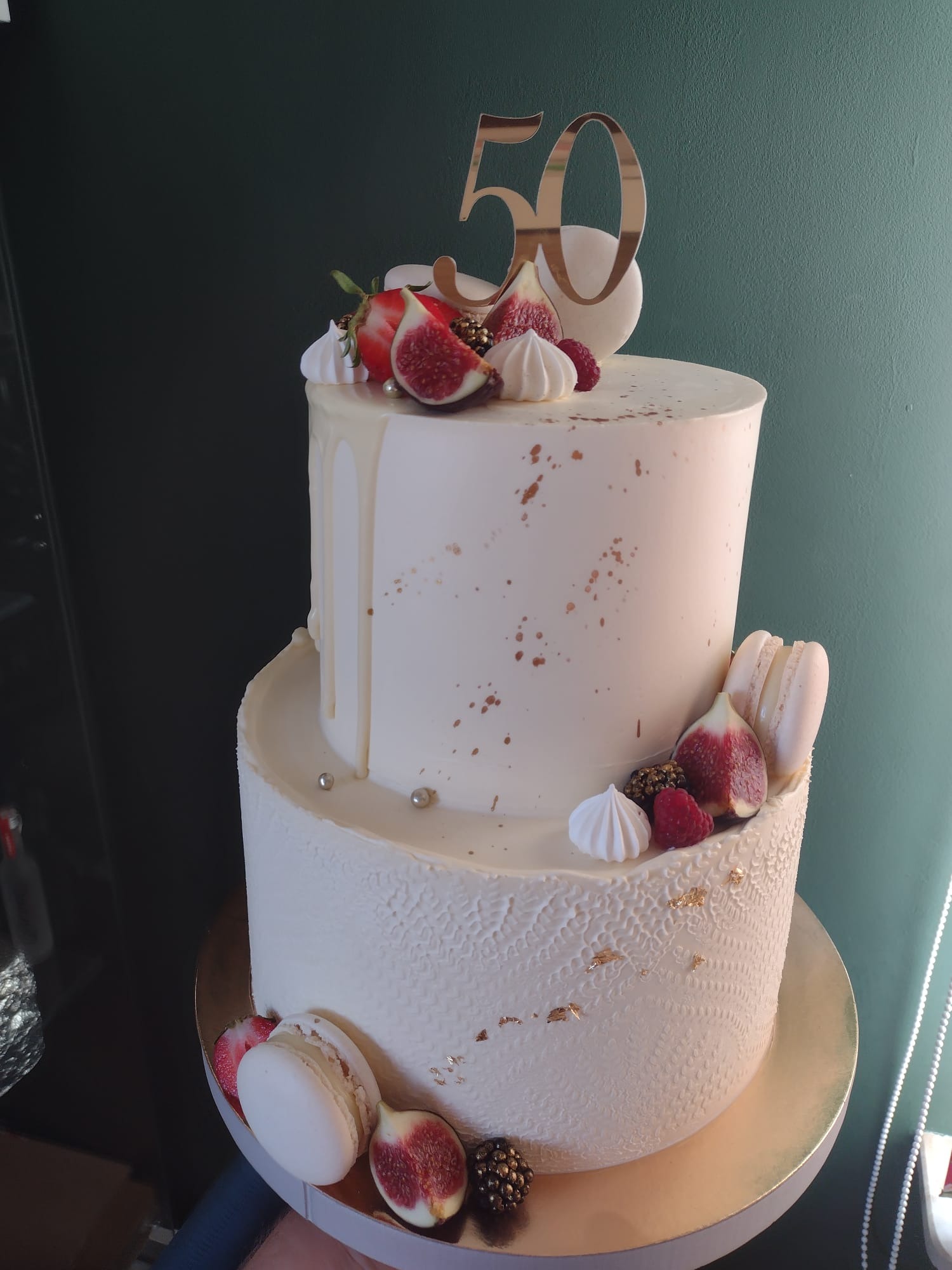 Dwupoziomowy tort urodzinowy w białej polewie, udekorowany figami, malinami, truskawkami, bezami i makaronikami, z topperem w kolorze złotym z liczbą 50.