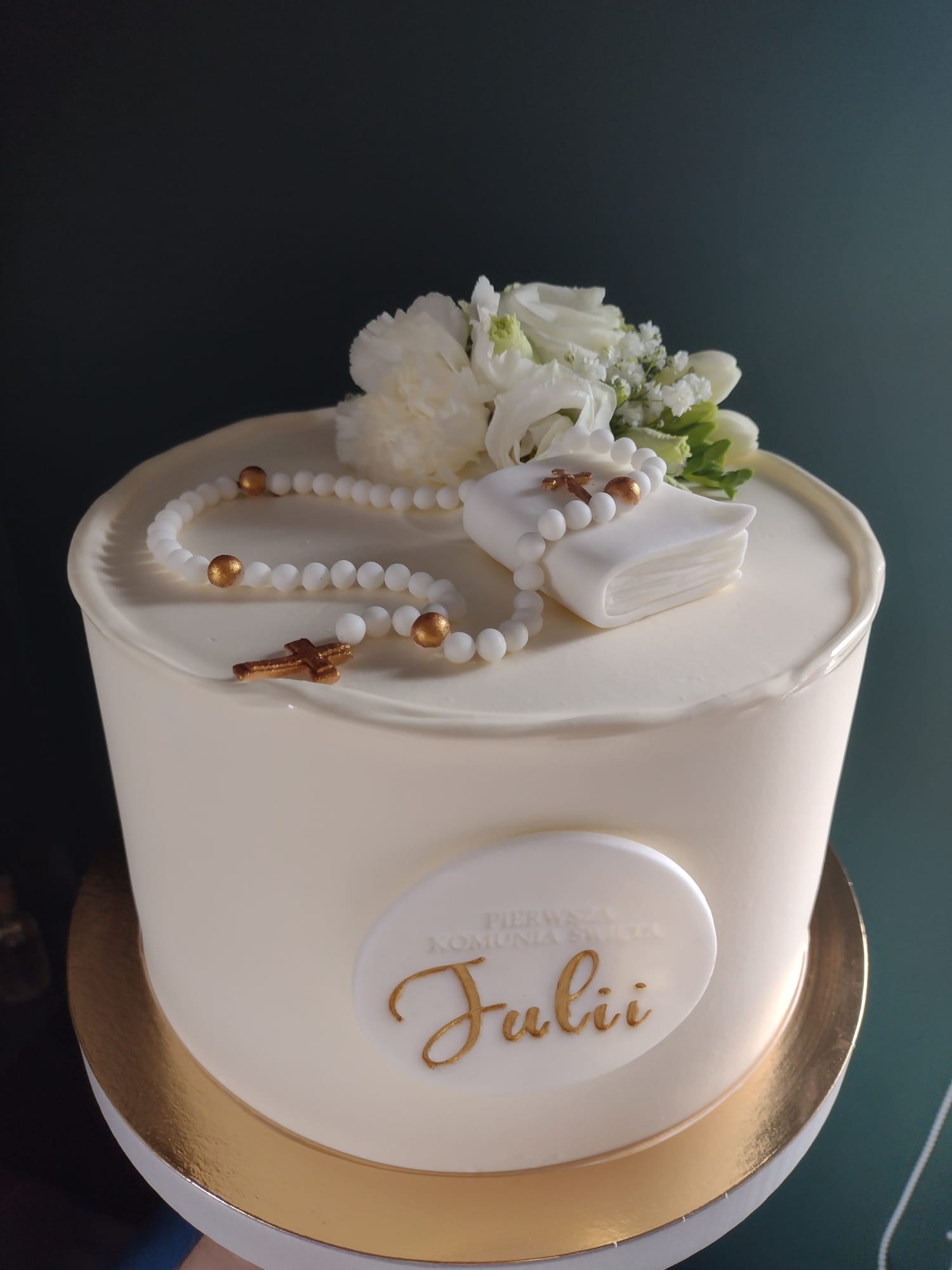 Tort komunijny z białym kremem, dekorowany różańcem z białych koralików i złotymi akcentami, małą białą książeczką oraz kwiatami. Na torcie umieszczono owalną plakietkę z napisem 'Pierwsza Komunia...