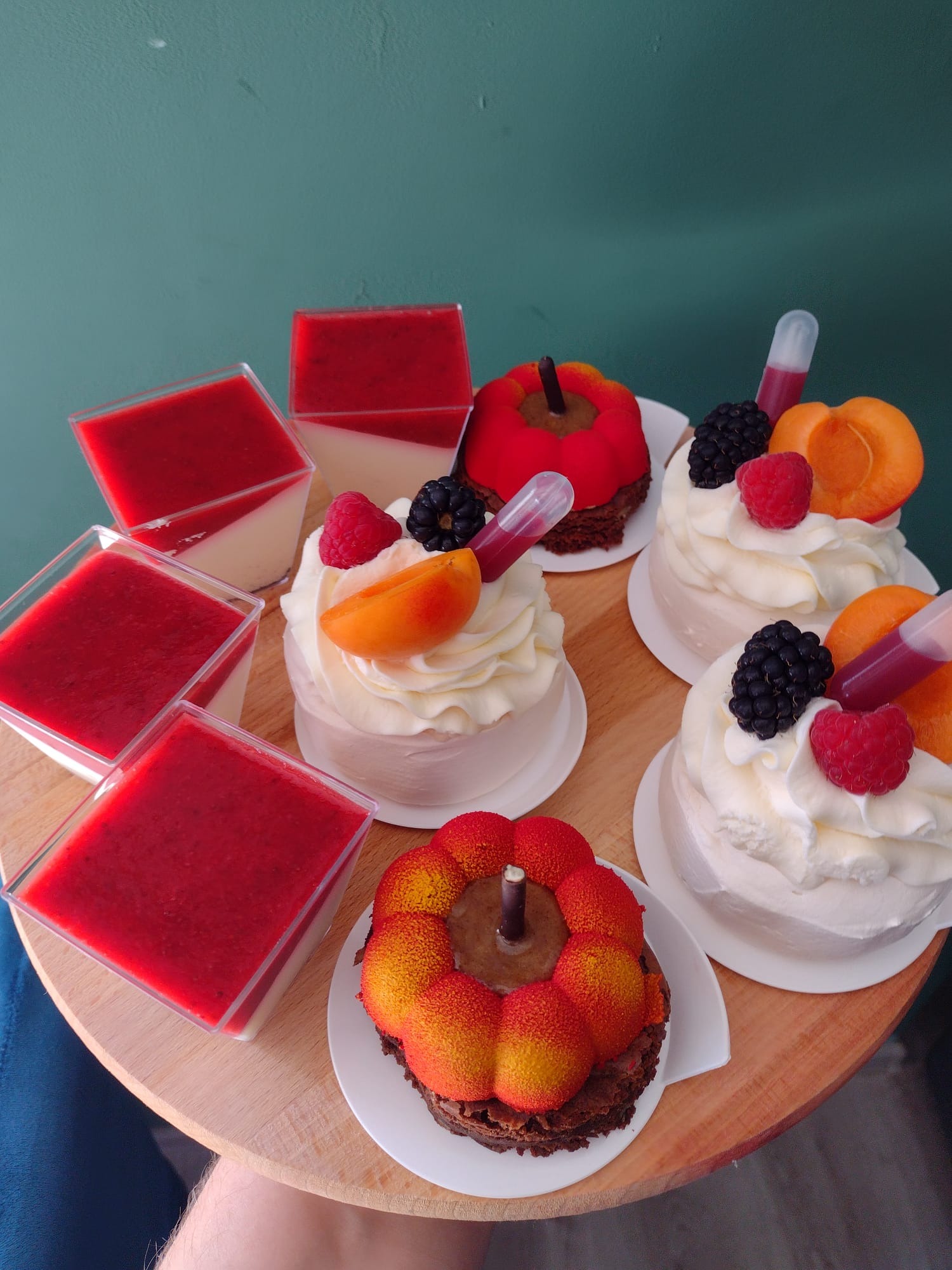 Zestaw mini deserów na drewnianej tacy: kremowe desery w kwadratowych naczyniach, mini torty z kremem, owocami (maliny, jeżyny, morele) i pipetą z sosem, oraz ciastka w kształcie jabłek z brązowym...