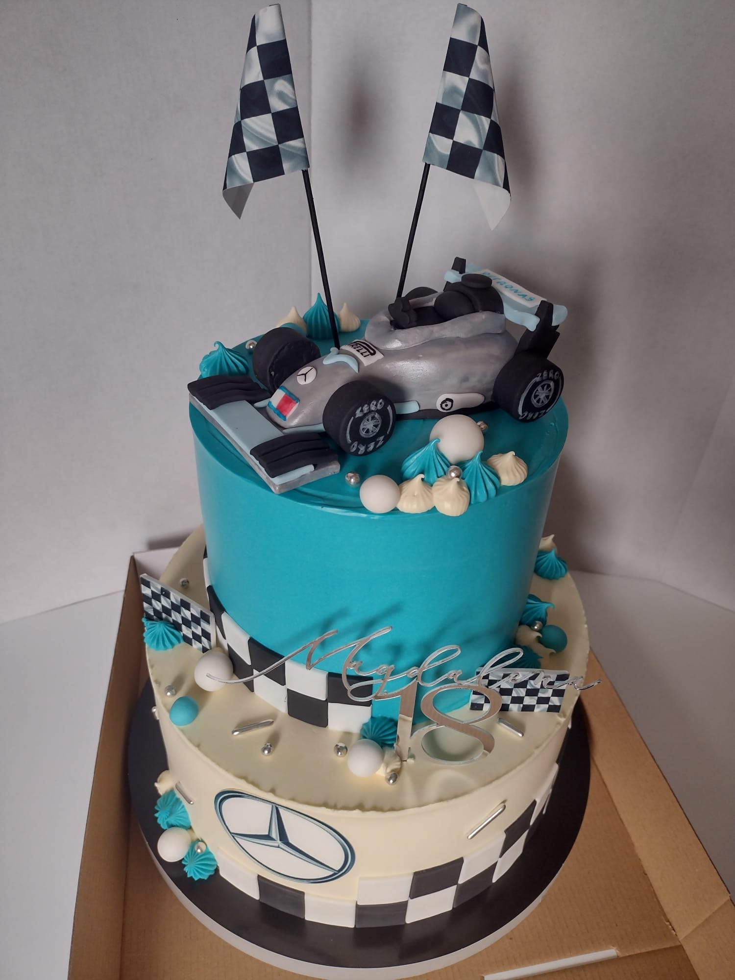 Dwupoziomowy tort urodzinowy z motywem Formuły 1: turkusowy górny poziom z modelem srebrnego bolidu i flagami w szachownicę, dolny poziom w kolorze kremowym z logo Mercedesa i dekoracjami...