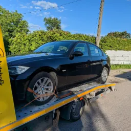 Czarny samoch&oacute;d osobowy marki BMW zabezpieczony pasami transportowymi na platformie lawety z ż&oacute;łtym panelem bocznym z logo firmy świadczącej usługi transportowe.