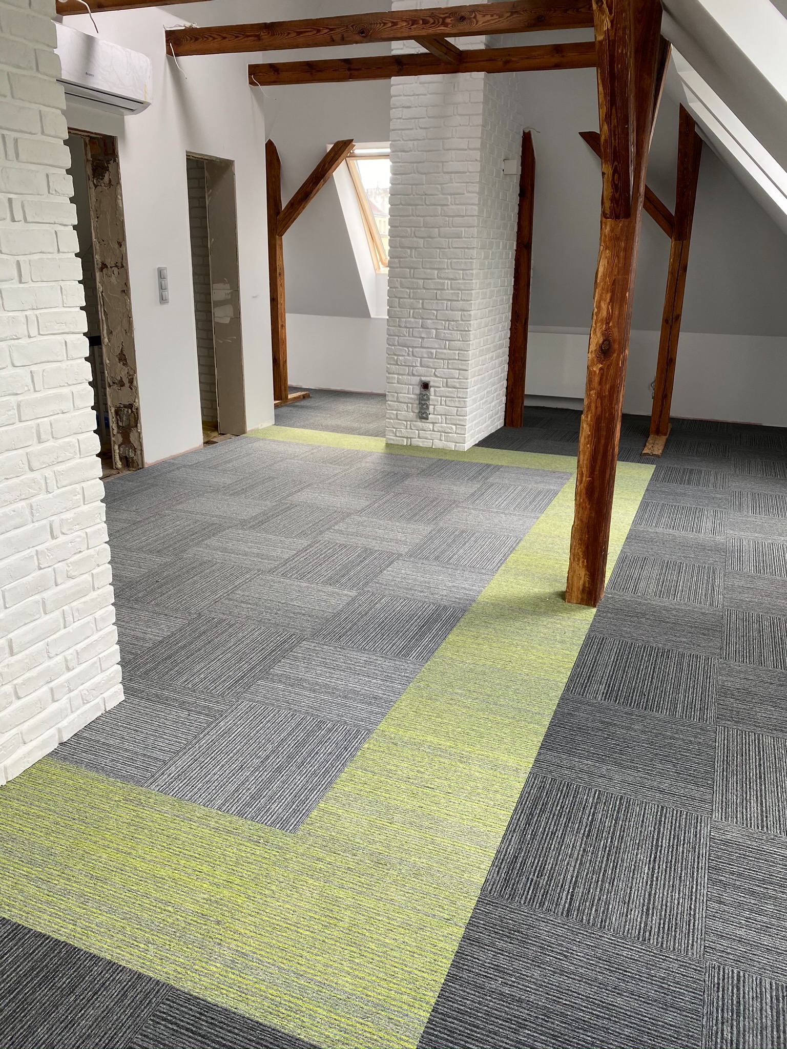 Wnętrze poddasza z szarą wykładziną w kwadraty z zielonym akcentem, drewniane belki konstrukcyjne, biała ceglana ściana i okno dachowe.