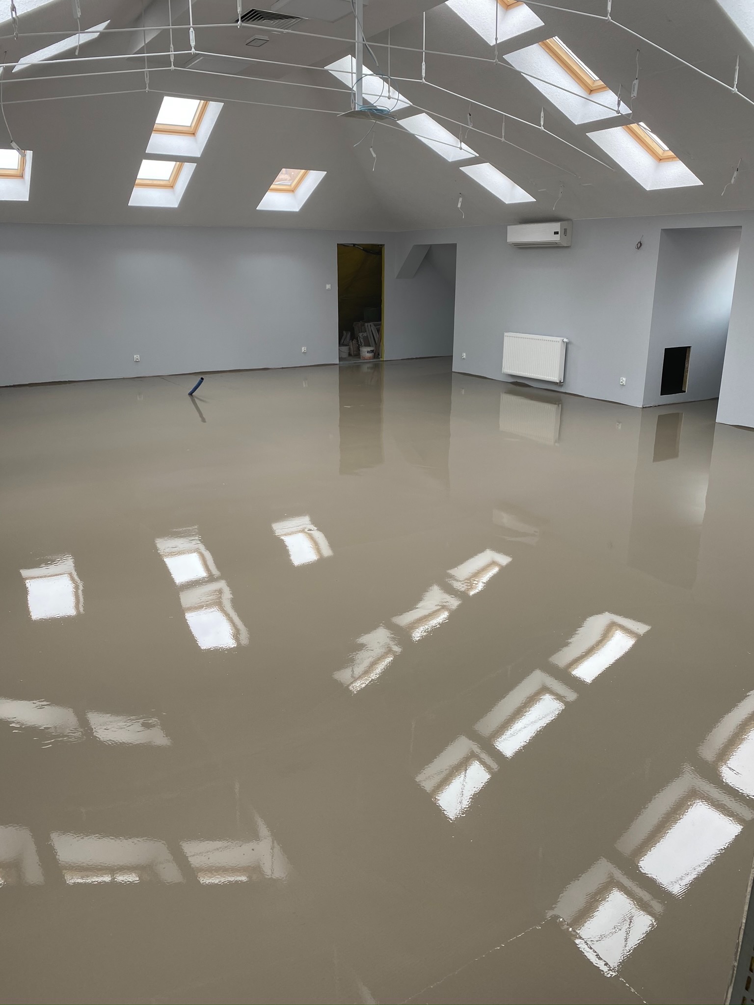 Wnętrze z wylewką samopoziomującą w odcieniach beżu, odbijającą światło z okien dachowych. Widoczne instalacje pod sufitem, grzejnik i klimatyzator na szarej ścianie.