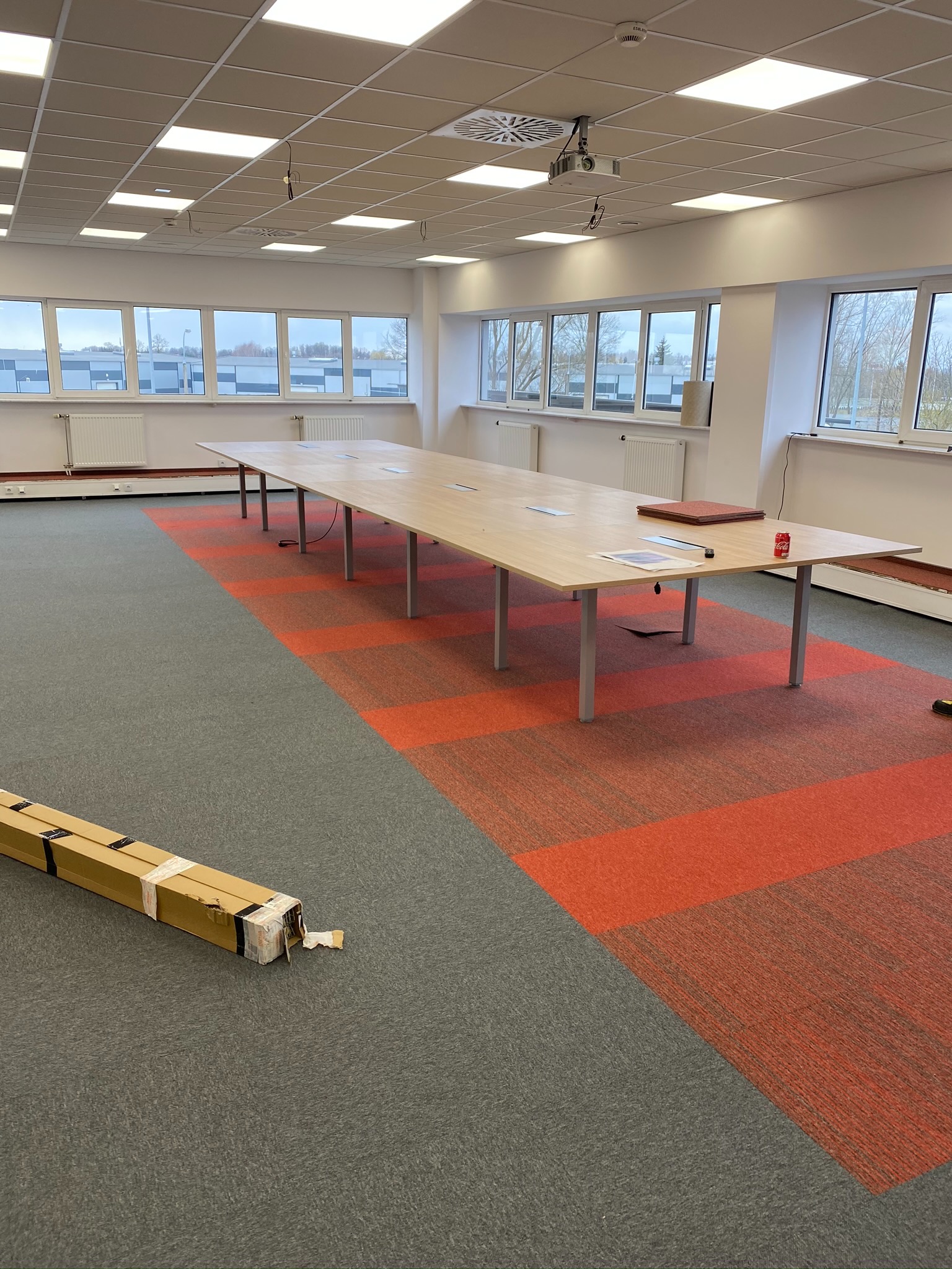 Wnętrze biura z długim stołem i częściowo położoną wykładziną dywanową w odcieniach szarości i czerwieni, widoczne opakowanie po wykładzinie, przestronne pomieszczenie z oknami.