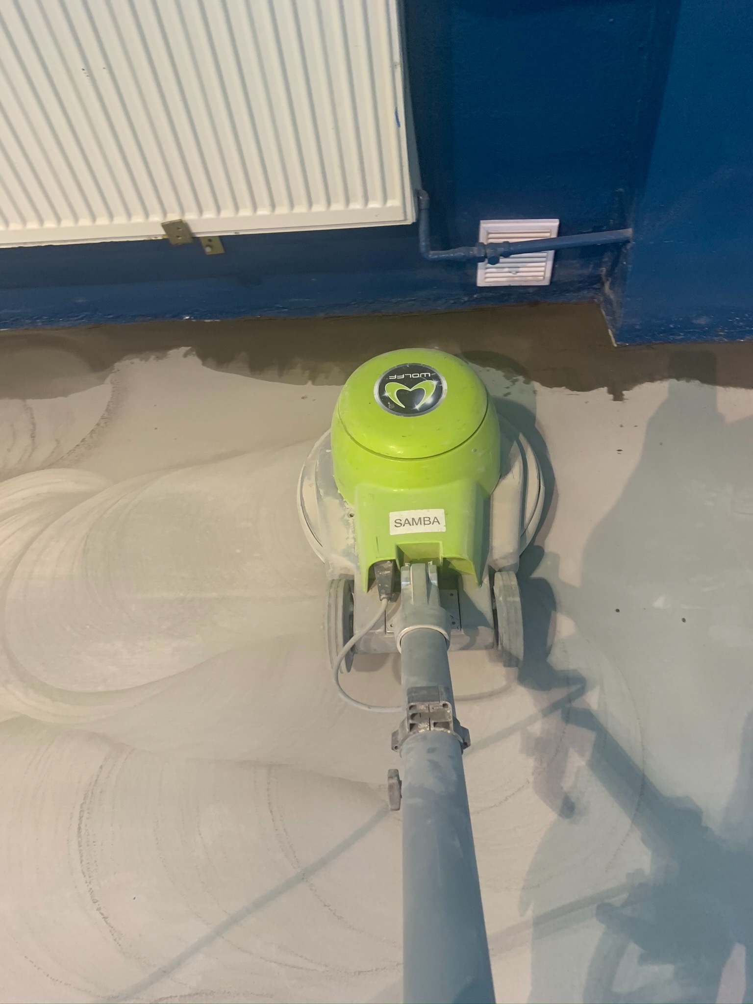 Zielona maszyna polerska marki Wolff, model Samba, używana do czyszczenia podłogi w pomieszczeniu z niebieskimi ścianami i grzejnikiem. Widoczne ślady po polerowaniu na szarej podłodze.