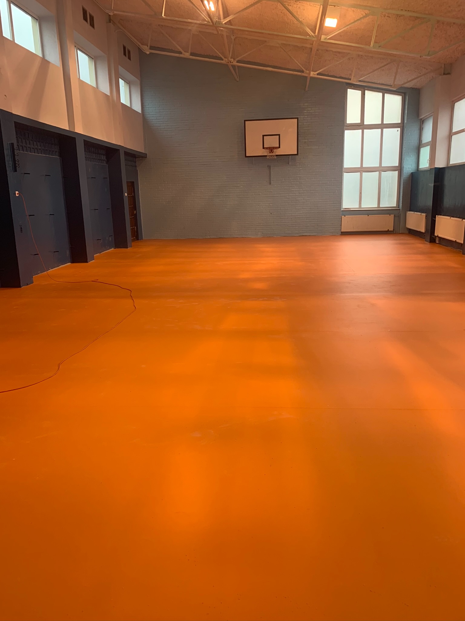 Pomarańczowa, poliuretanowa podłoga w hali sportowej z koszem do koszykówki na ścianie, widoczne okna i grzejniki.