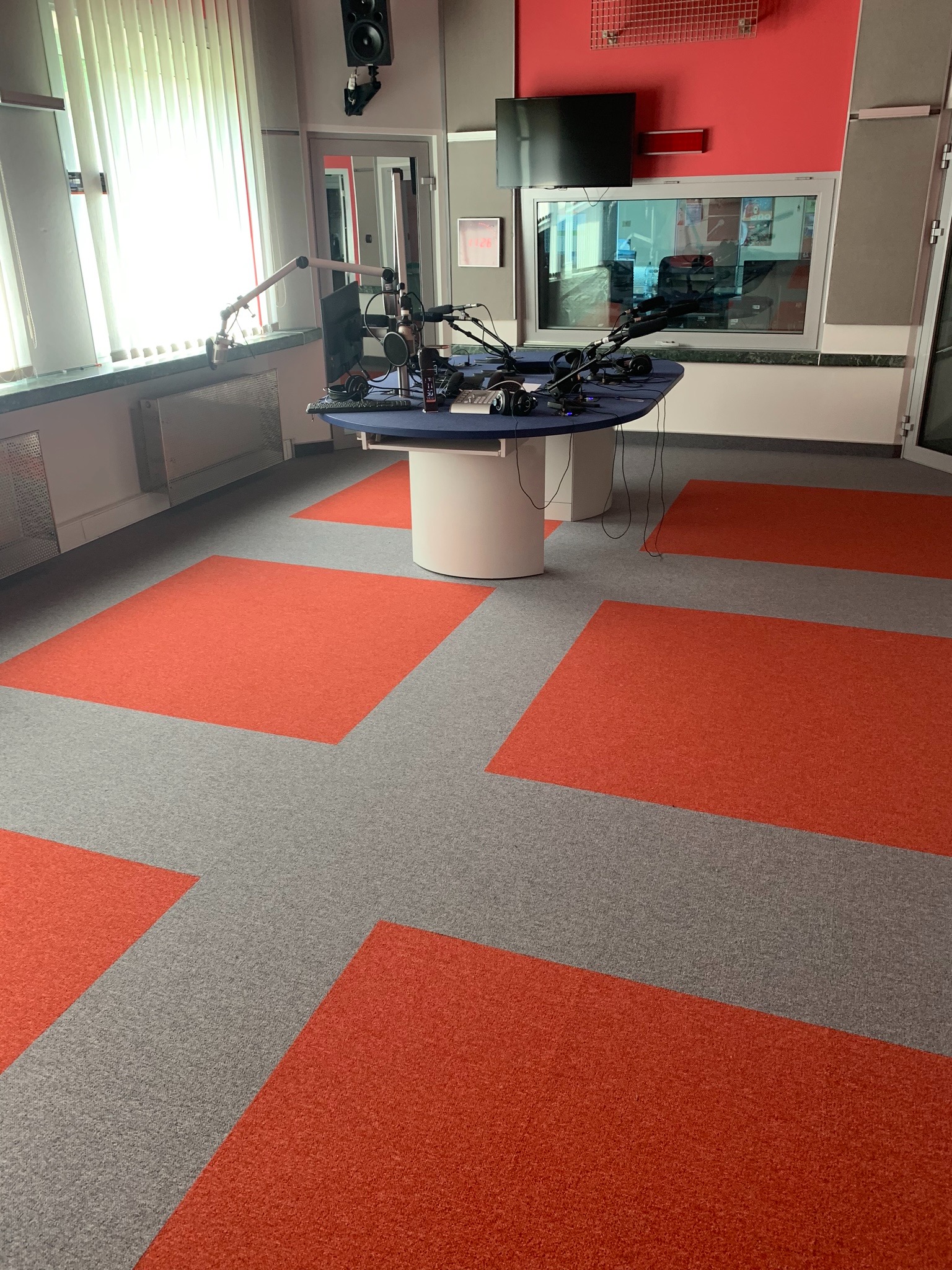 Studio radiowe z szarą wykładziną w kwadraty z pomarańczowymi akcentami, okrągły stół z mikrofonami i słuchawkami.