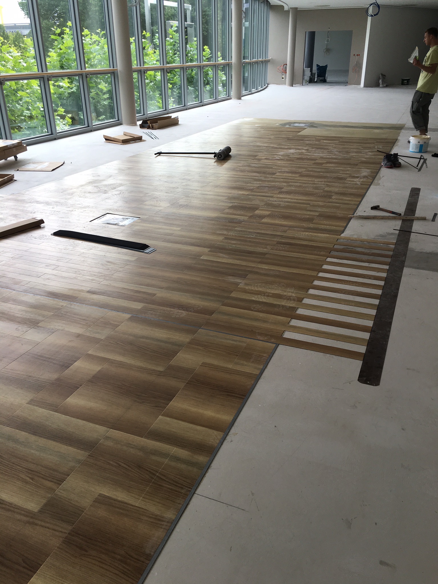 Układanie podłogi z paneli imitujących drewno w przestronnym pomieszczeniu z dużymi oknami, widoczne narzędzia i częściowo ułożona powierzchnia, pracownik w tle.